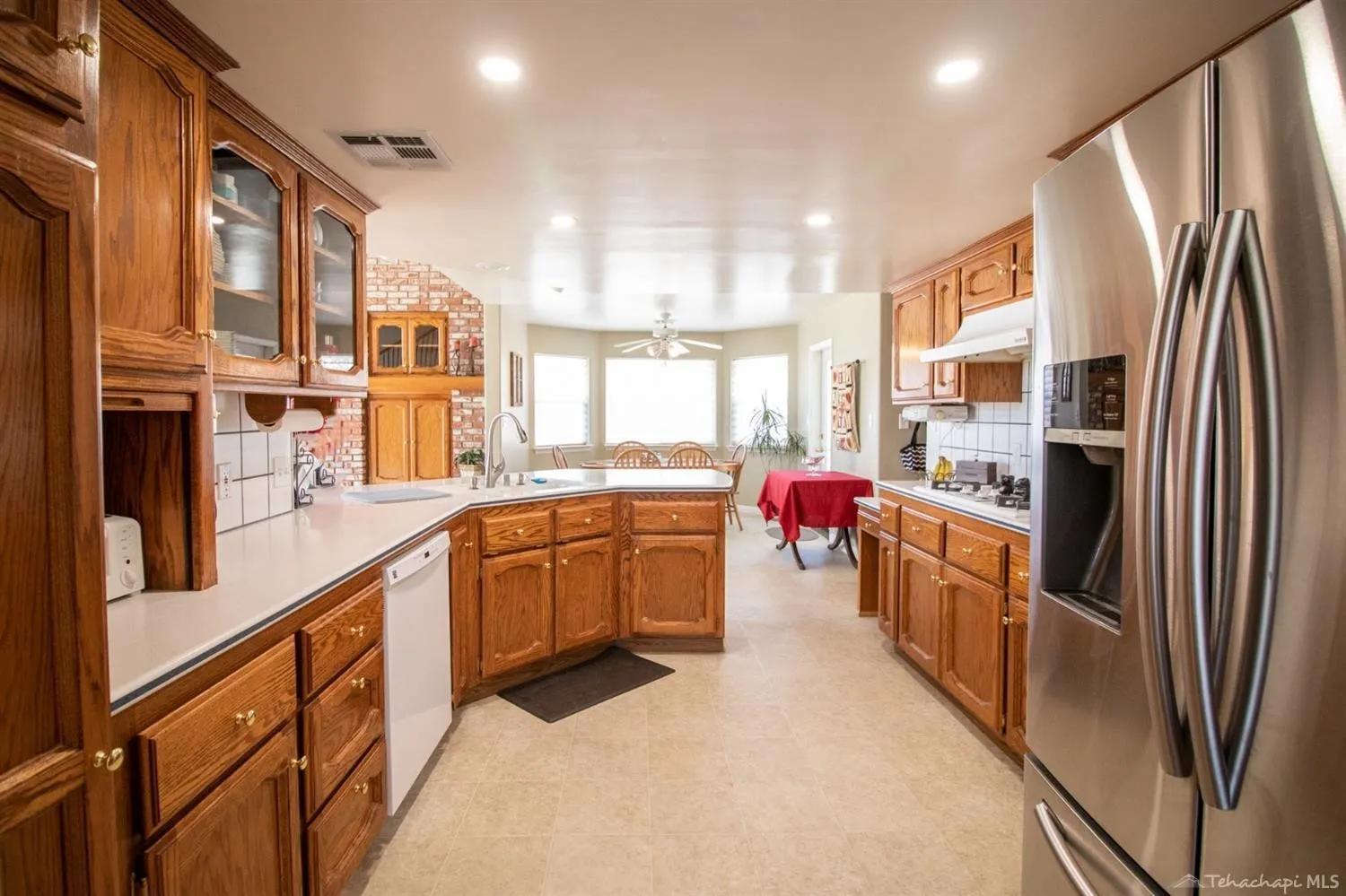 Opposite view of the same oak kitchen showing stainless fridge, white dishwasher, oak glass-front upper cabinets, and a brick accent wall near the breakfast nook with bay windows.