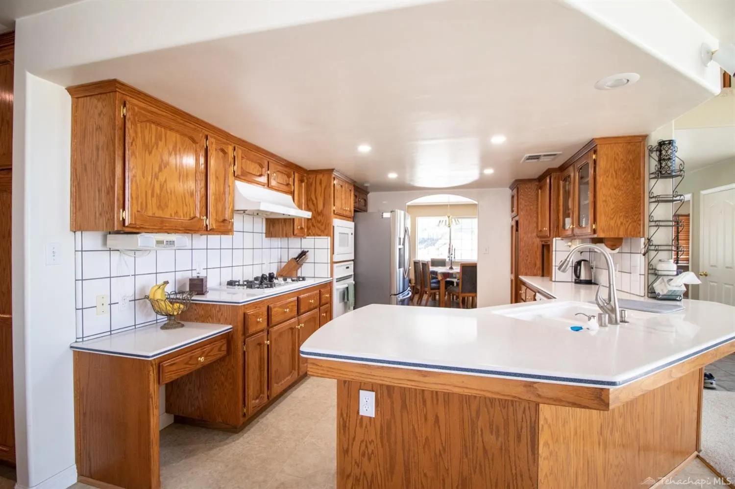 Original 1990s kitchen with orange oak raised-panel cabinets, white tile countertops with blue trim, speckled tile floor, and a built-in oak desk nook on the left wall.