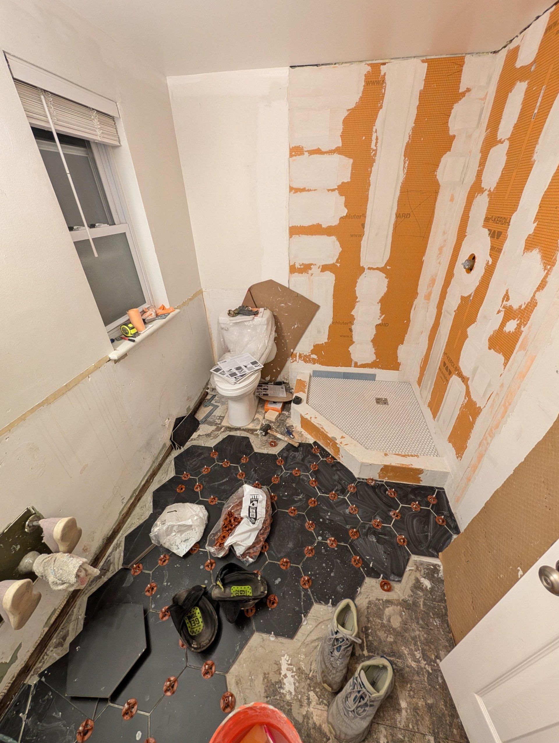 Bathroom mid-tile with orange Schluter Kerdi waterproofing board covering the shower walls, a white penny tile shower pan already in place, and large black hexagon floor tiles set with orange leveling clips, work boots and tile bags scattered around the floor.