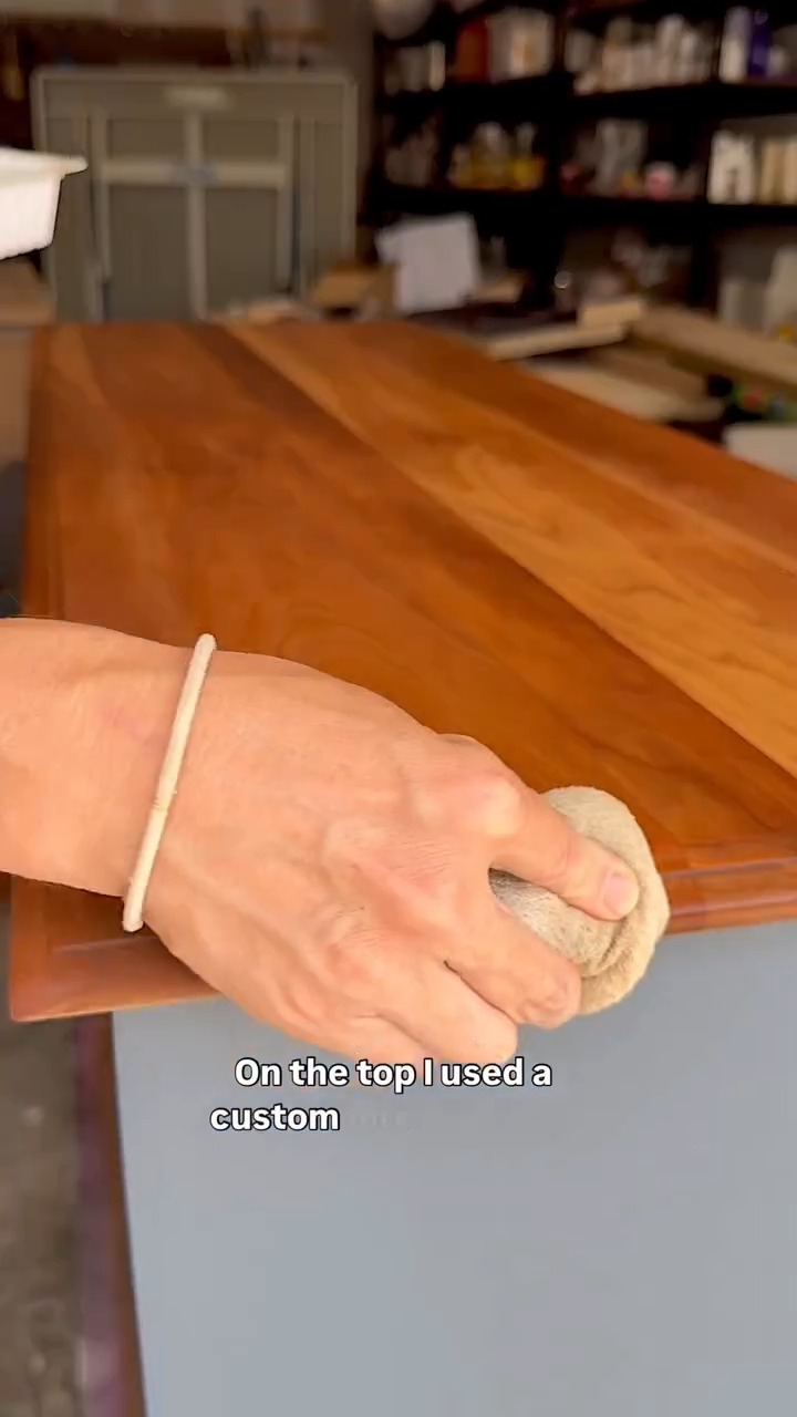 A hand with a bracelet rubbing a cloth across the raw wood dresser top, tinting the pale grain to a warm honey tone, with navy painted body visible below.