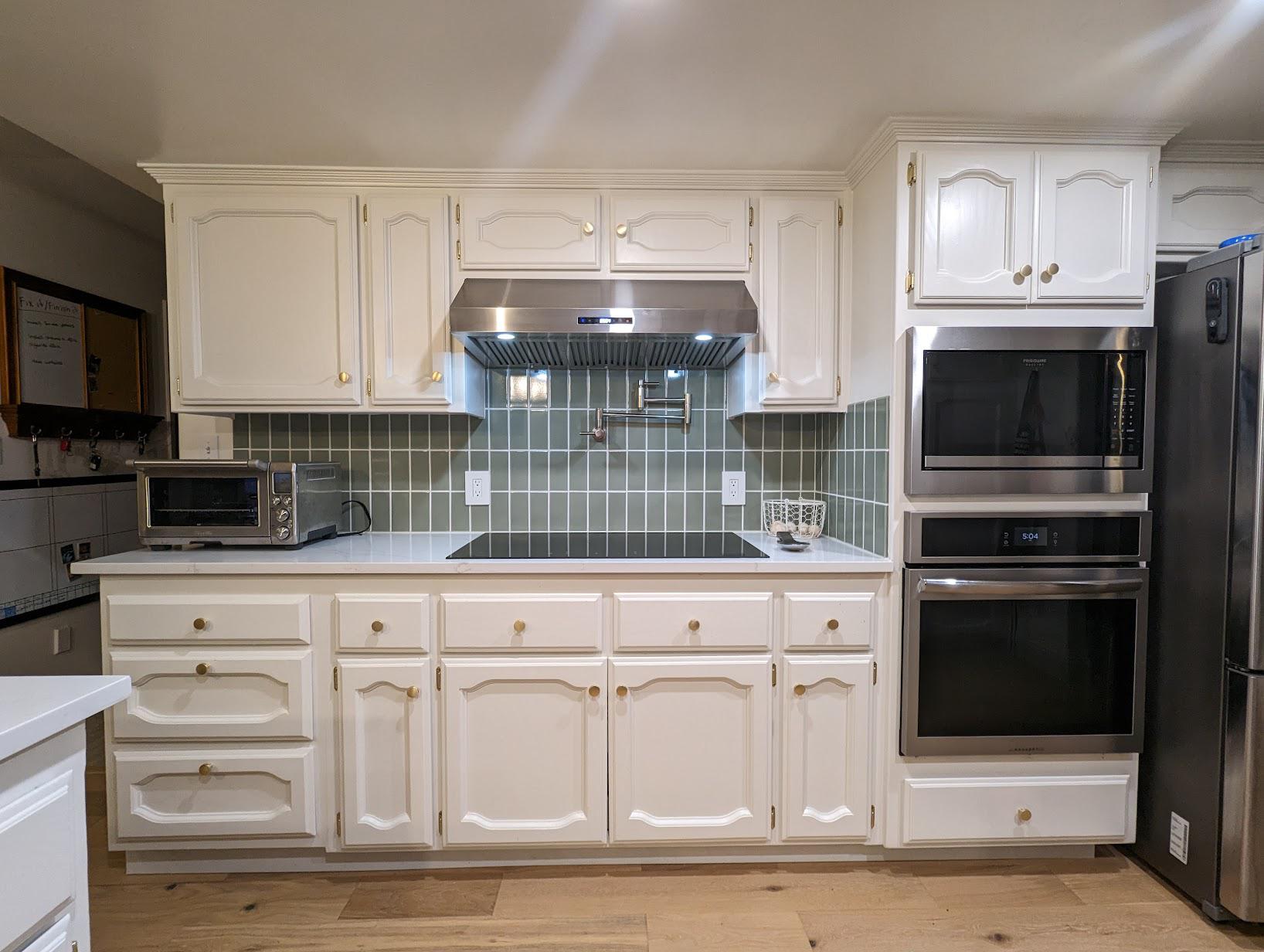 After view of the painted white cabinets with the same raised-panel profile, now paired with a sage green vertical stack tile backsplash, stainless hood, pot filler, induction cooktop, and brass knobs, with a stacked wall oven and microwave on the right.