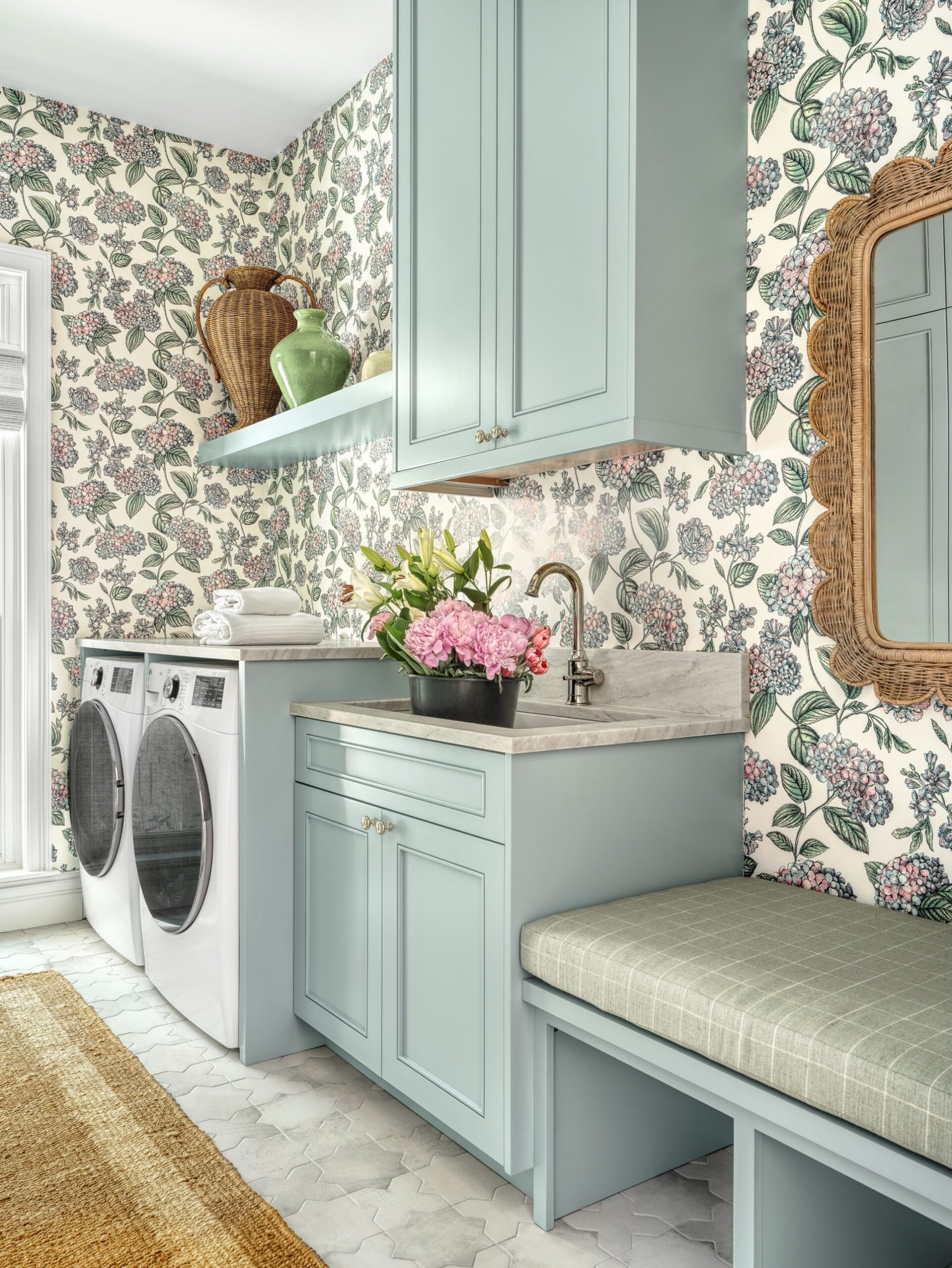 Pale blue cabinets paired with hydrangea floral wallpaper in a laundry mudroom with a side-by-side white washer and dryer, marble sink top with pink peonies, upholstered checked bench, and scalloped wicker mirror