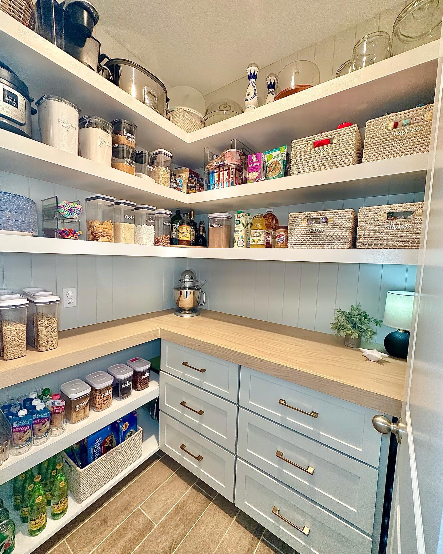 Pale blue beadboard pantry with L-shaped butcher block counters, white open corner shelves holding labeled clear canisters, wicker baskets, pale blue cabinets with brass pulls, and a small black lamp in the back corner