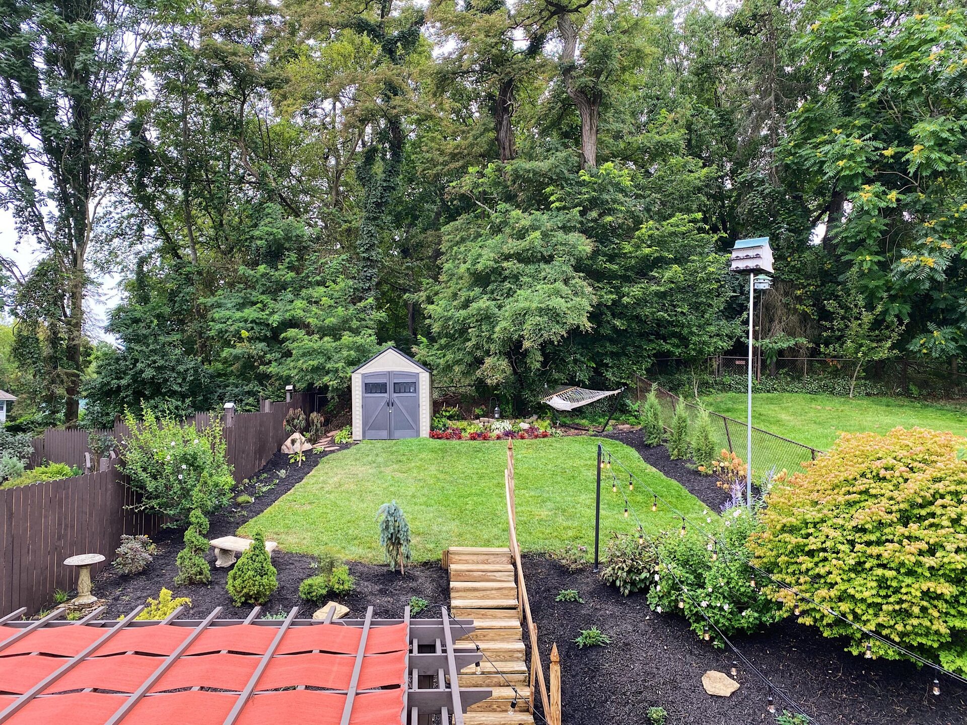 Dark brown pergola with red shade sail strips on the slats, Edison bulb string lights hanging below, wicker lounge chairs with orange cushions and striped pillows, and a mulched slope with new wooden stairs behind it.