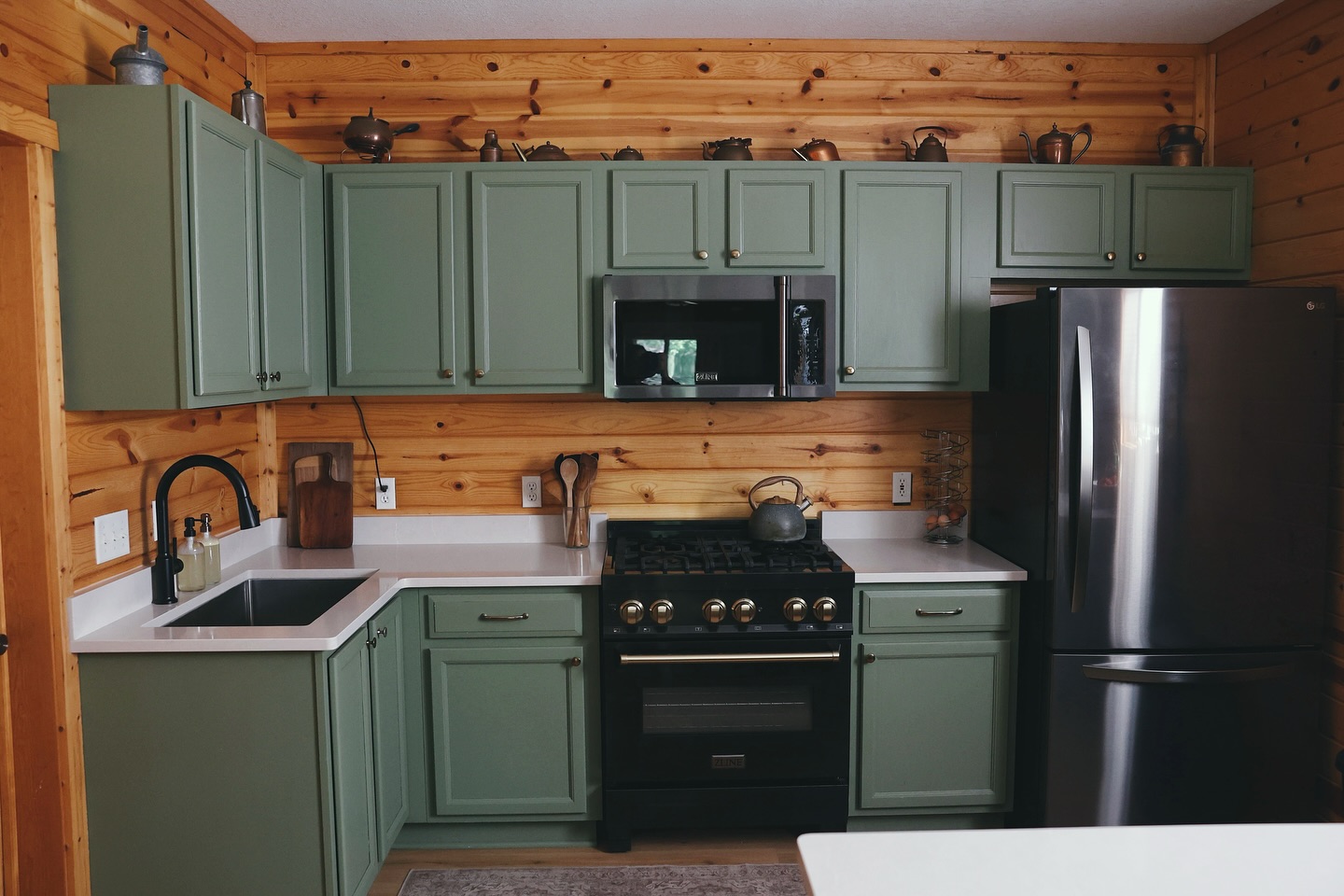Small cabin kitchen with sage green raised-panel cabinets, horizontal knotty pine wall paneling, a black gas range with brass knobs, black stainless refrigerator, and antique copper teapots and kettles lined up along the top of the upper cabinets