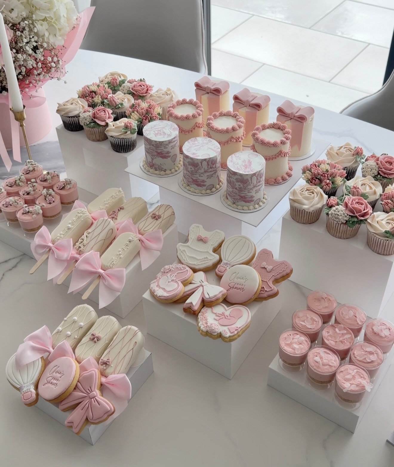 Pink and cream dessert table featuring bow-topped cakes, piped rose cupcakes, pink toile printed mini cakes, and iced hot air balloon cookies on white risers