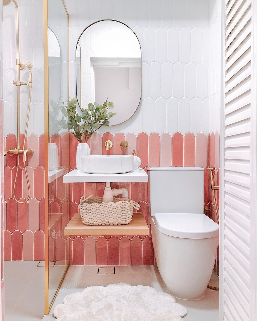 Small bathroom with pink fish-scale wall tiles on the lower half and white fish-scale on the upper half, floating white shelf with a wood lower shelf, scalloped white vessel sink, oval brass-rimmed mirror and brass exposed-pipe shower fixtures