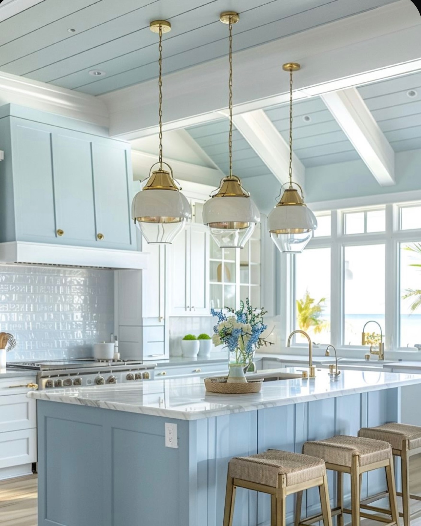 Coastal kitchen with soft powder blue painted cabinets and a matching shiplap ceiling with white beams, three brass pendants with white dome shades over a white marble island, pale blue tile backsplash, and a palm tree view through the windows