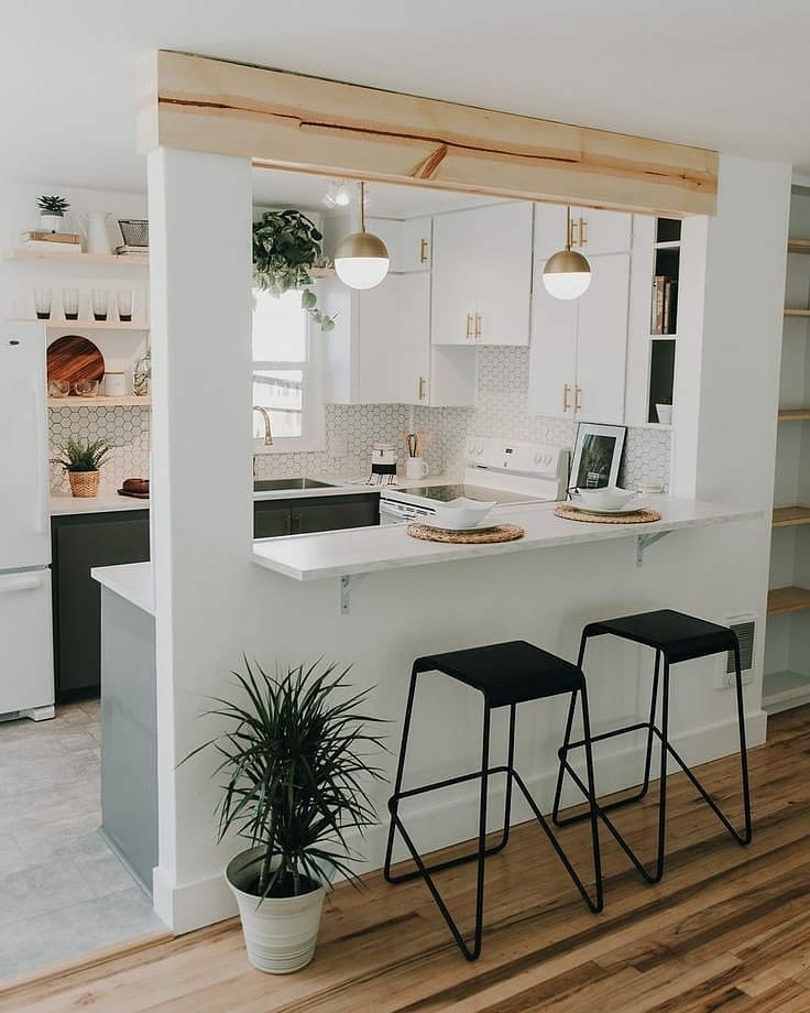 Small open kitchen with a raw wood beam above the opening, white cabinets with brass hardware, white hex tile backsplash, two black bar stools, and a floor plant