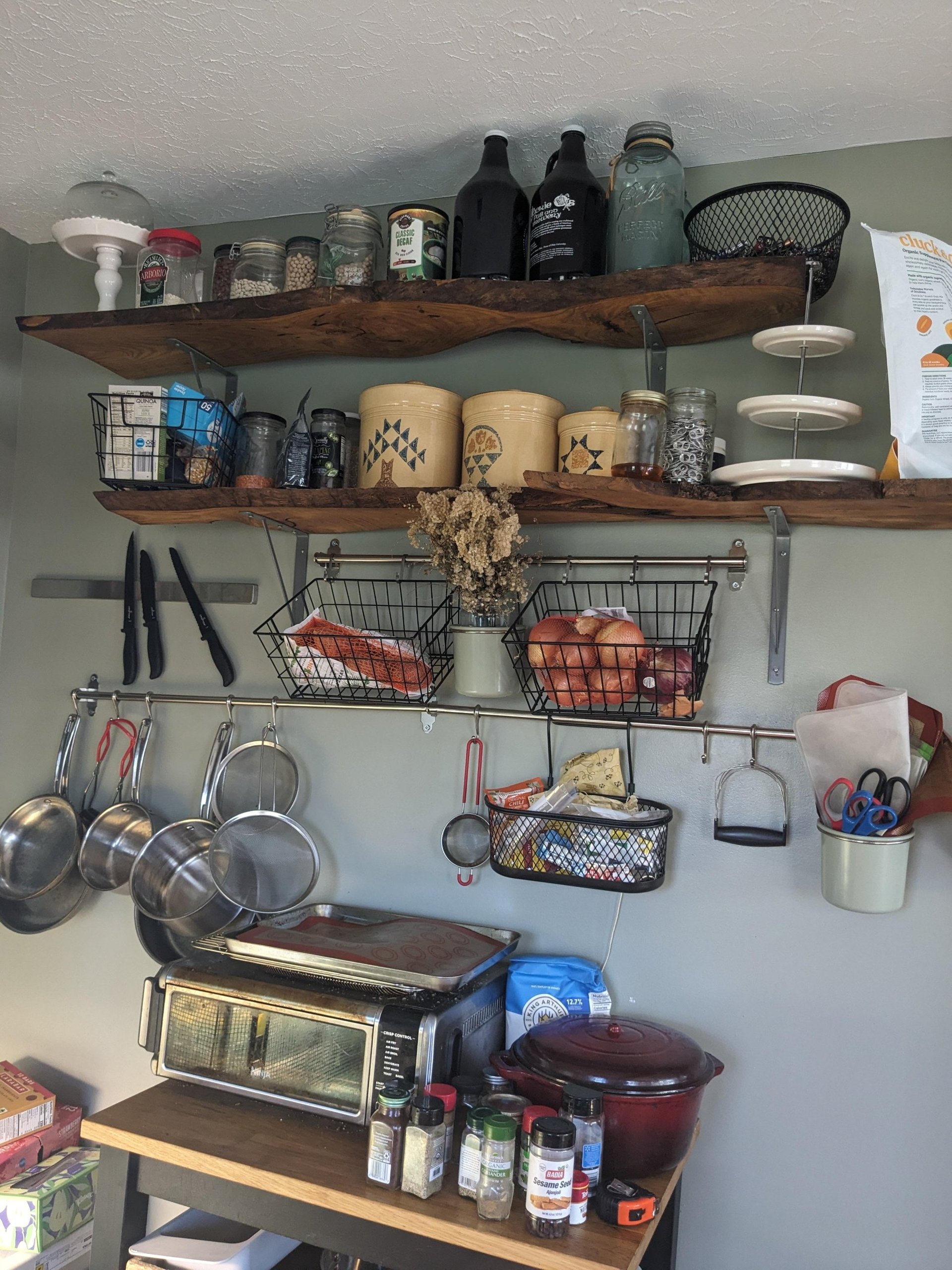 Before kitchen back wall with two rough live-edge wood shelves nailed up as makeshift storage, holding jars of beans and quinoa, with pots hanging from a rail below.