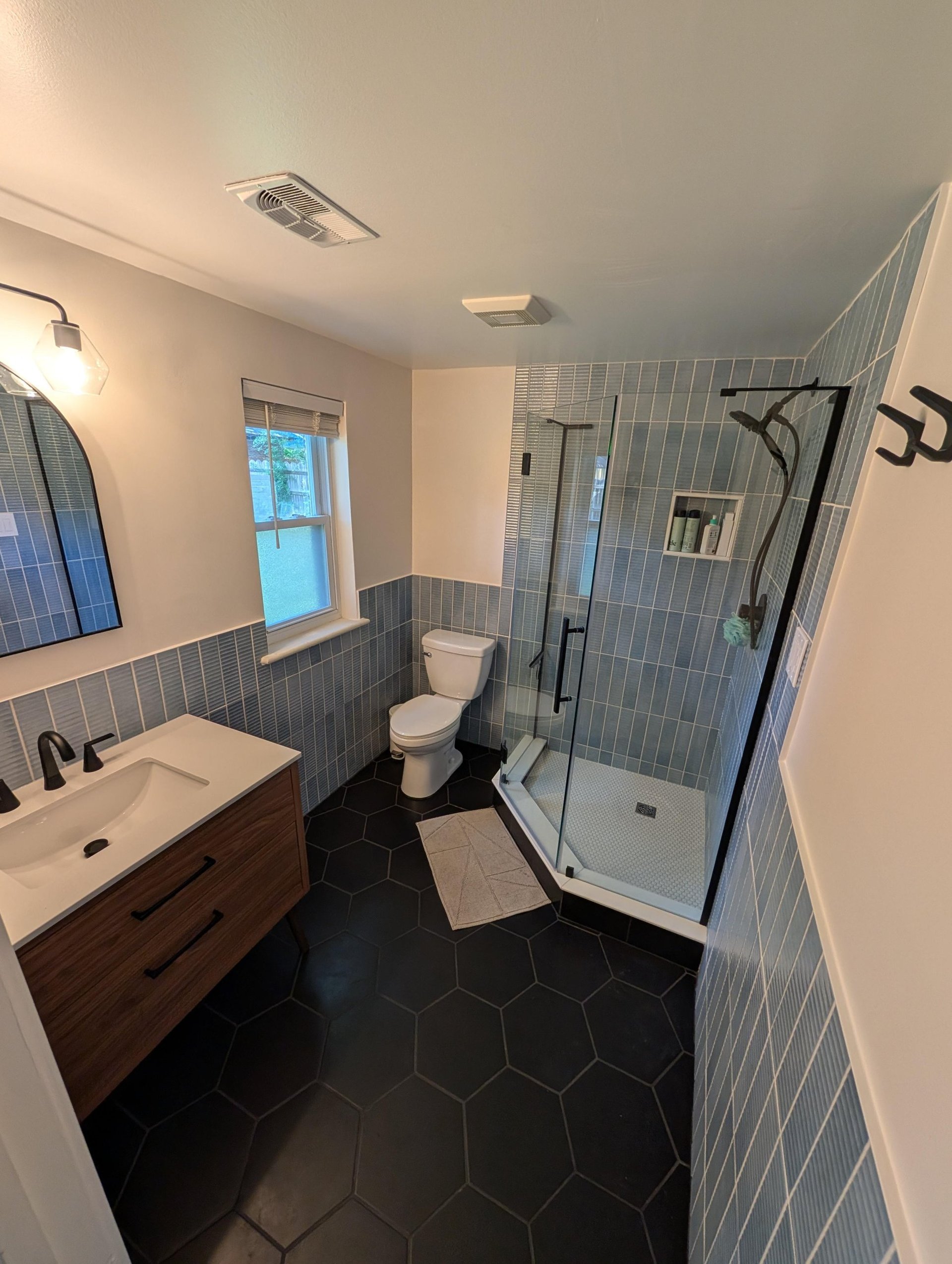 Finished bathroom with dusty blue vertical stacked wall tile running as wainscoting around the room, a walnut floating vanity with a white quartz top, black matte fixtures, an arched black mirror, large black hexagon floor tile, and a frameless glass neo-angle shower in the far corner.