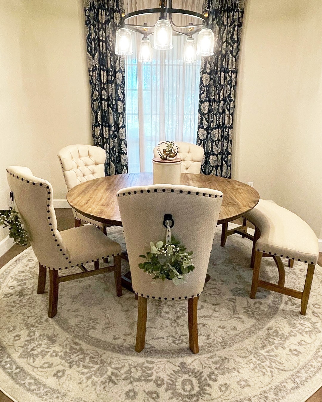 : Round farmhouse dining nook with wood table, cream upholstered dining chairs with black nailhead trim, a curved upholstered bench, dark blue and white floral curtains, and a mason jar chandelier overhead