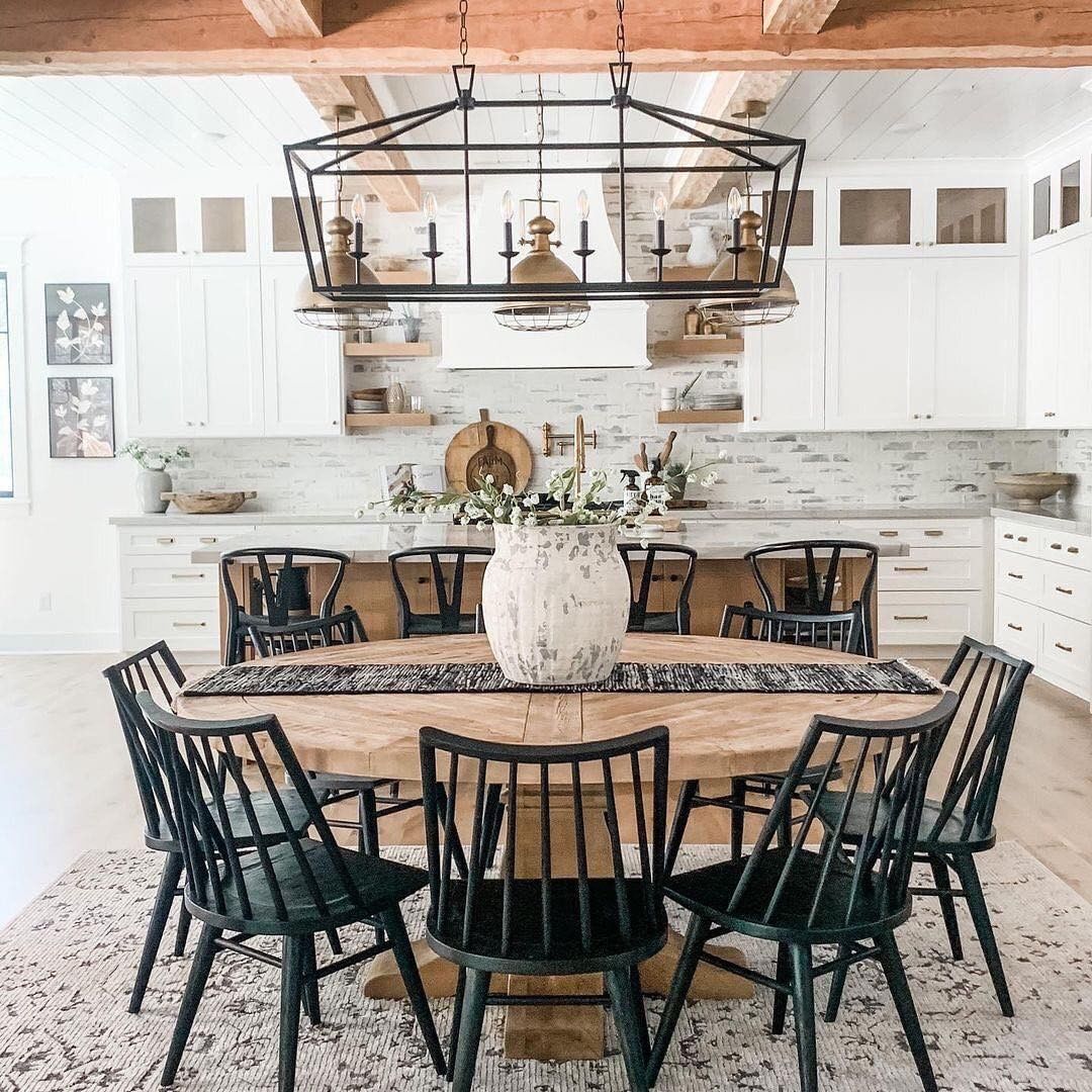 Modern farmhouse dining with large round chunky wood table, ring of black spindle Windsor chairs, large iron lantern chandelier, white kitchen with wood ceiling beams and gray brick backsplash in background