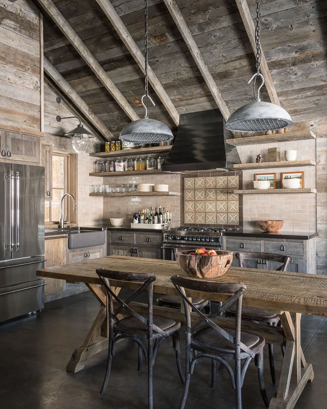 Rustic cabin kitchen with weathered gray wood cabinets, vaulted pine ceiling with heavy beams, two zinc dome pendants on chains, farmhouse sink, open floating shelves with glass jars, patterned tile behind the range, and an X-base reclaimed wood dining table with cross-back chairs