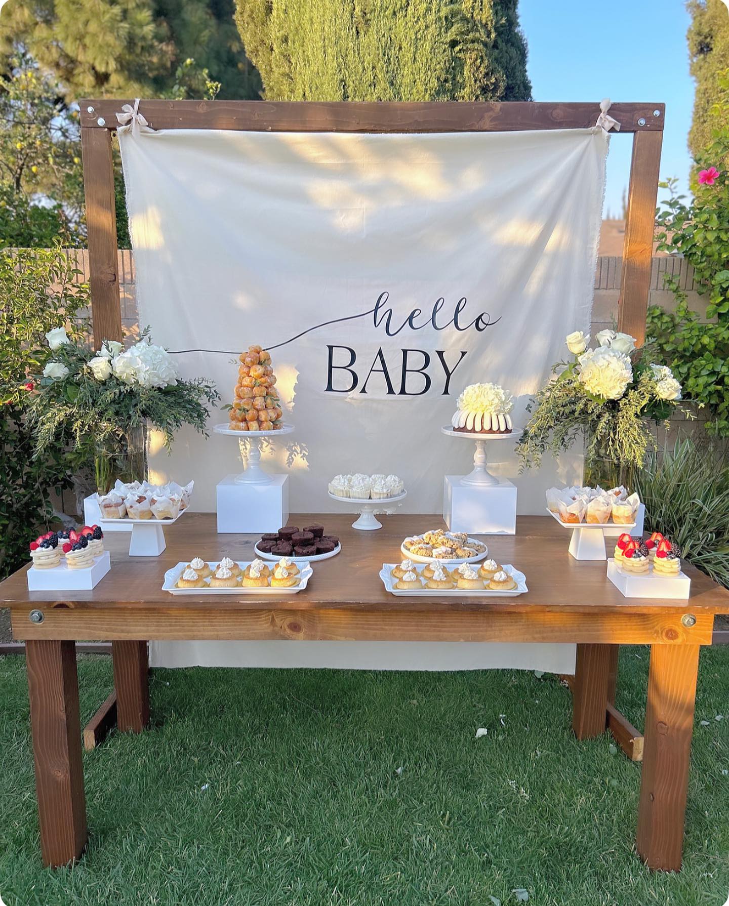 Rustic outdoor hello baby dessert table on a wood farm table with a cream fabric backdrop, croquembouche tower, bundt cake with flowers, berry pavlovas, and brownies