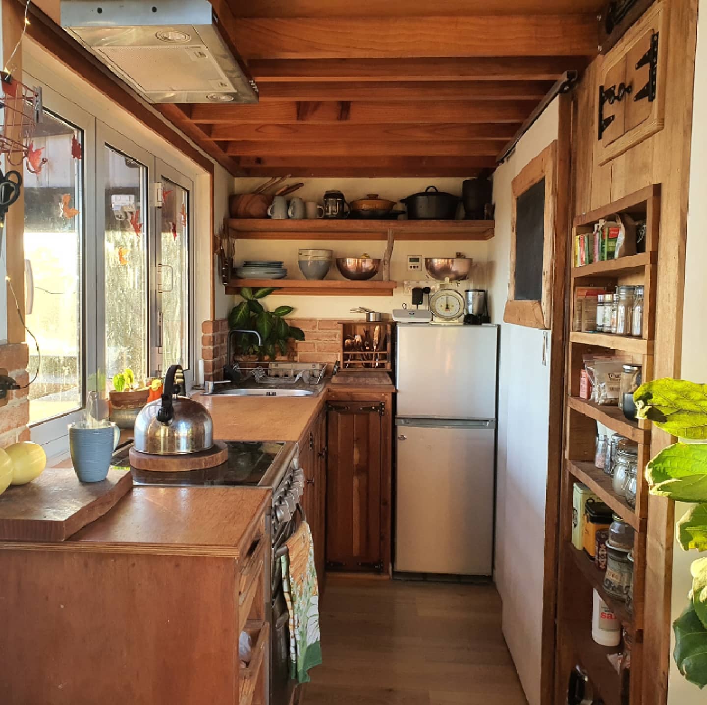 Rustic wood tiny house kitchen with brick backsplash, open wood shelves holding copper bowls and ceramic mugs, a chalkboard, a vintage scale, a stainless kettle on the stove, and a tall wood spice rack