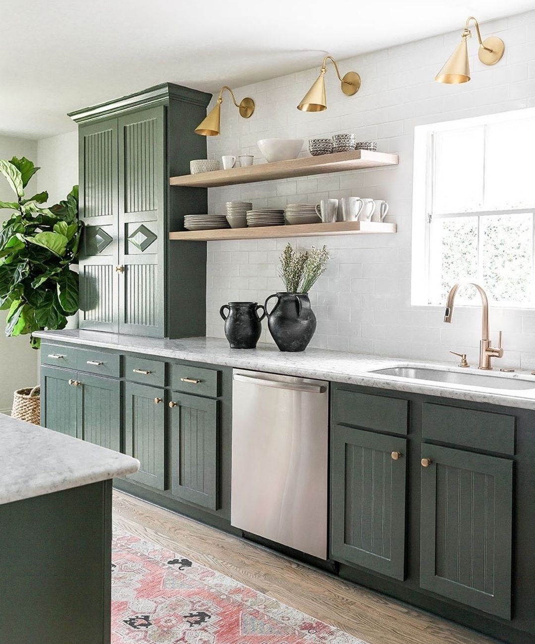 Muted sage green beadboard kitchen cabinets with brass knobs and bar pulls, two brass cone sconces above white oak floating shelves, a white subway tile backsplash, white marble countertops, a champagne brass faucet, and a fiddle leaf fig in the corner