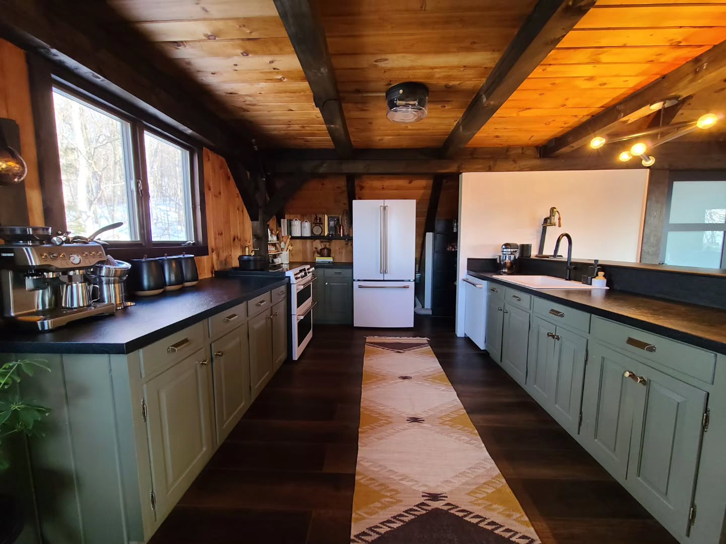 Galley-style cabin kitchen with sage green shaker cabinets, black countertops, dark exposed wood beams on a pine ceiling, white double oven range, white French door refrigerator, and a woven cream and brown kilim runner on dark wood floors