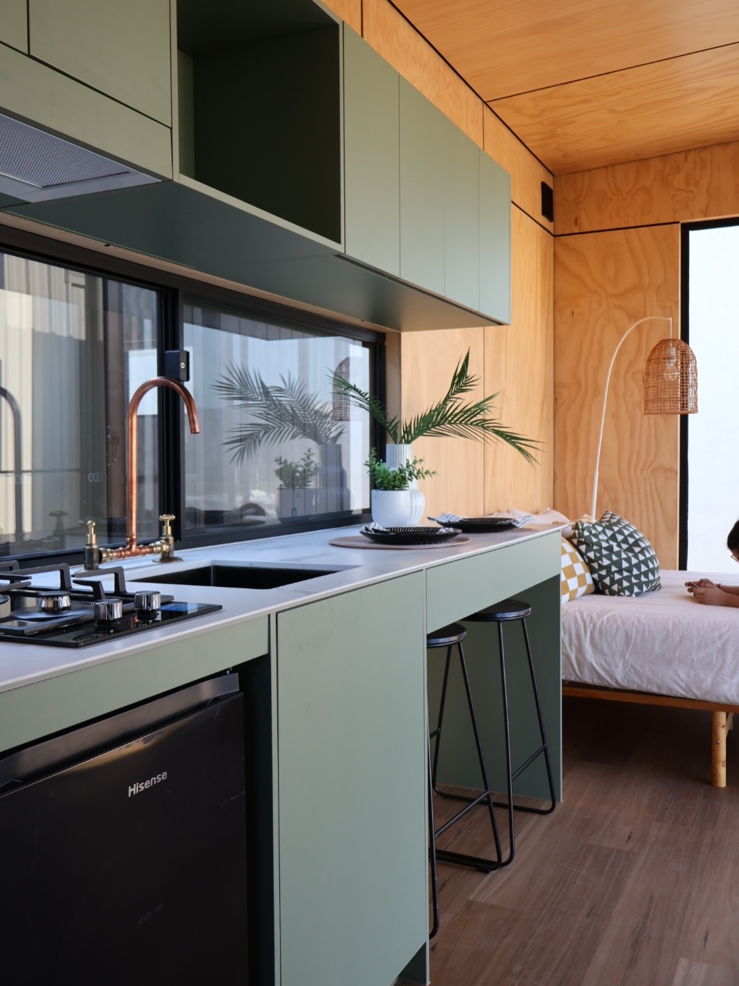 Sage green tiny house kitchen with white marble counter, copper gooseneck faucet, matte black gas cooktop, plywood walls, and a bed with patterned pillows visible in the adjoining room