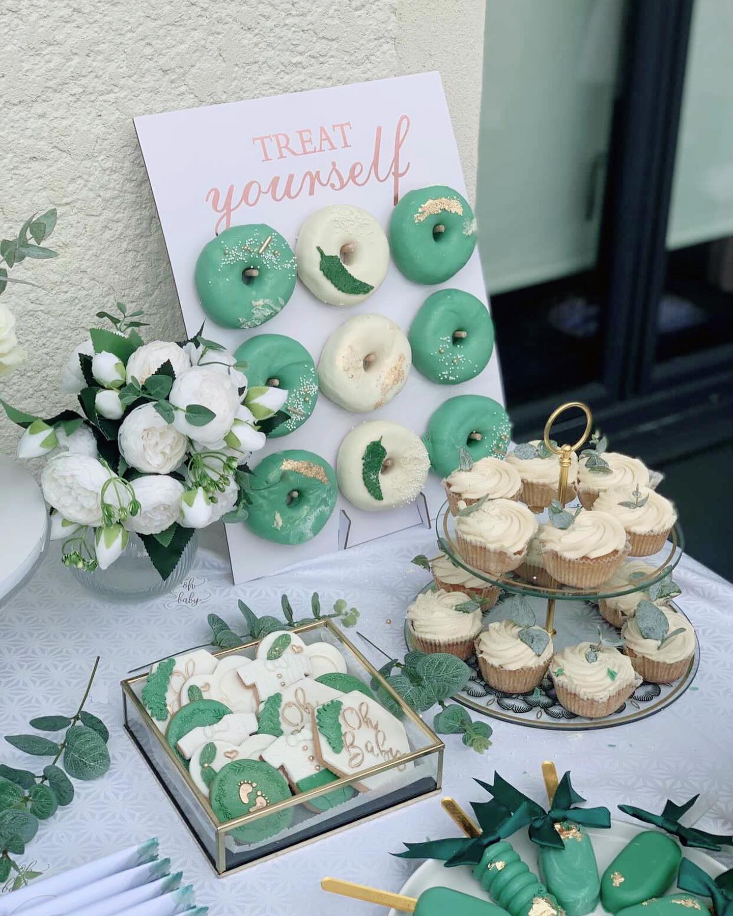 Sage green and white donut wall with treat yourself sign, white peony flower arrangement, cream cupcakes with eucalyptus, and oh baby iced cookies in a glass box