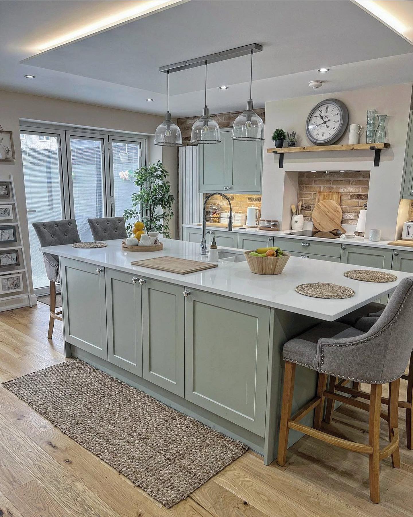 Sage green kitchen with a long island, three glass dome pendants, a reclaimed brick alcove with a wood mantel shelf, and gray tufted bar chairs with nailhead trim