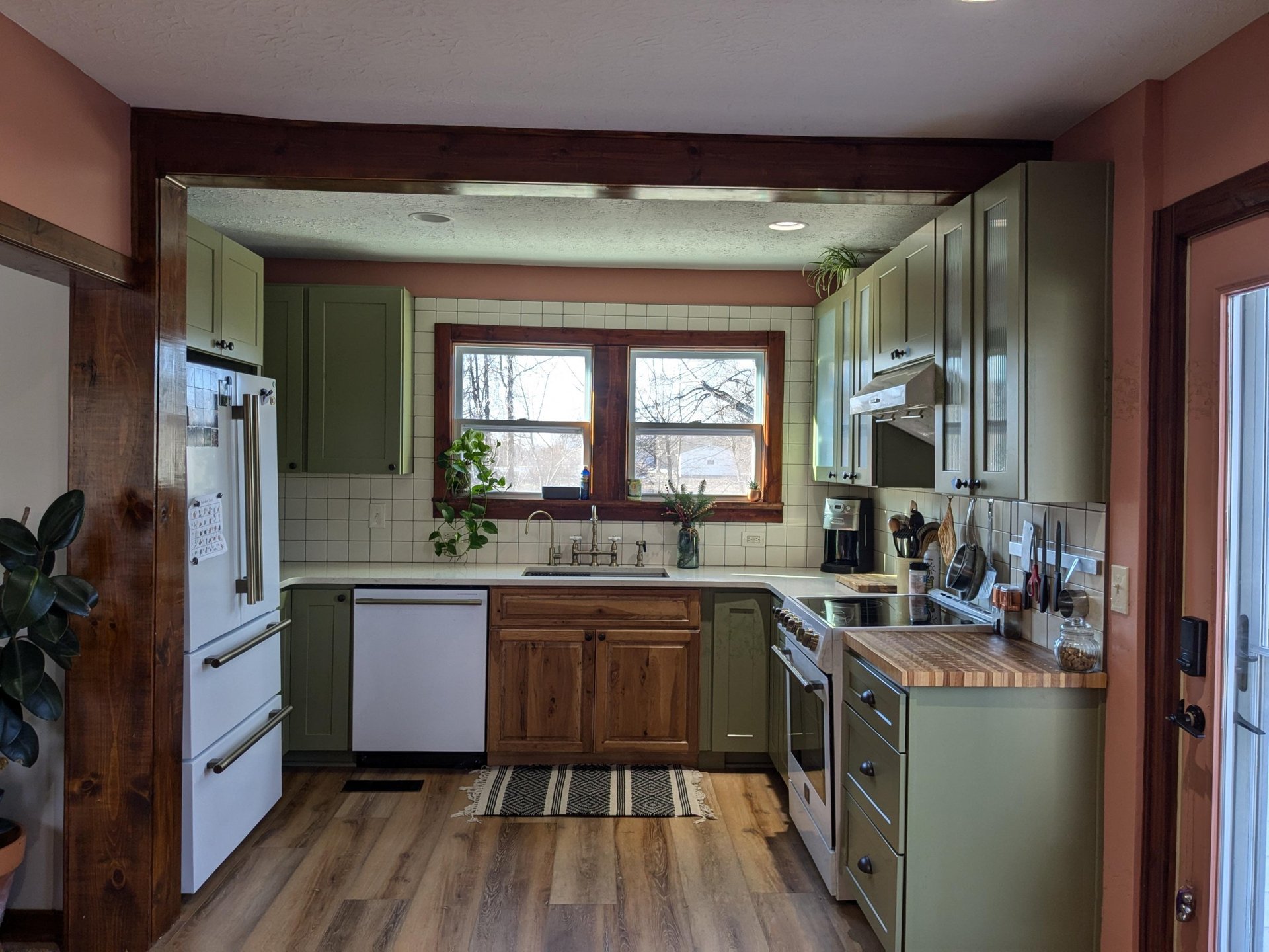 Wide view of the finished kitchen with sage green shaker cabinets, coral pink walls, white square tile backsplash, a brass bridge faucet, a natural hickory sink cabinet, and white quartz counters.