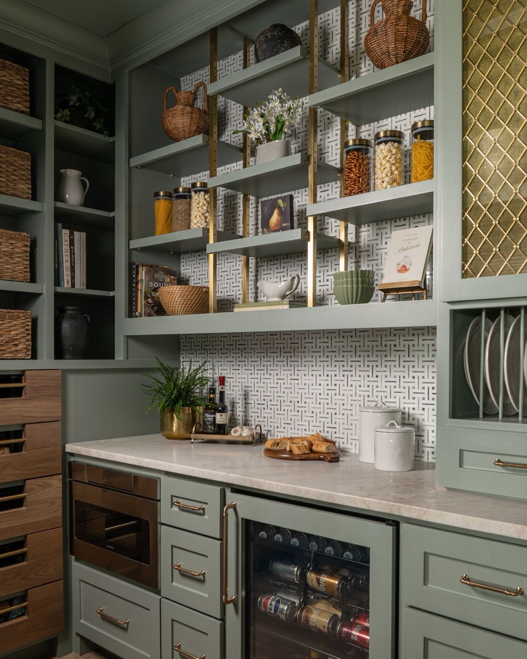 Sage green painted pantry cabinets with gold mesh wire cabinet door, patterned white and black tile backsplash, built-in beverage fridge, marble counter, and vertical dish plate rack