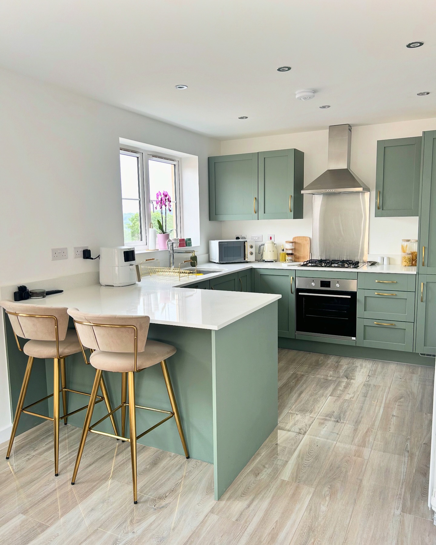 Sage green shaker kitchen in a U-shape with gold bar pull hardware, white quartz counters, a stainless chimney hood, and blush pink velvet barstools on gold legs