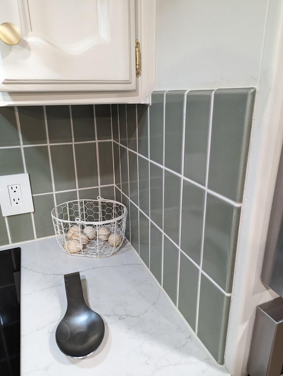 Close-up of the sage green vertical tile backsplash meeting the white quartz counter with soft gray veining, a wire basket of eggs on the counter, and a hint of the painted upper cabinet above.