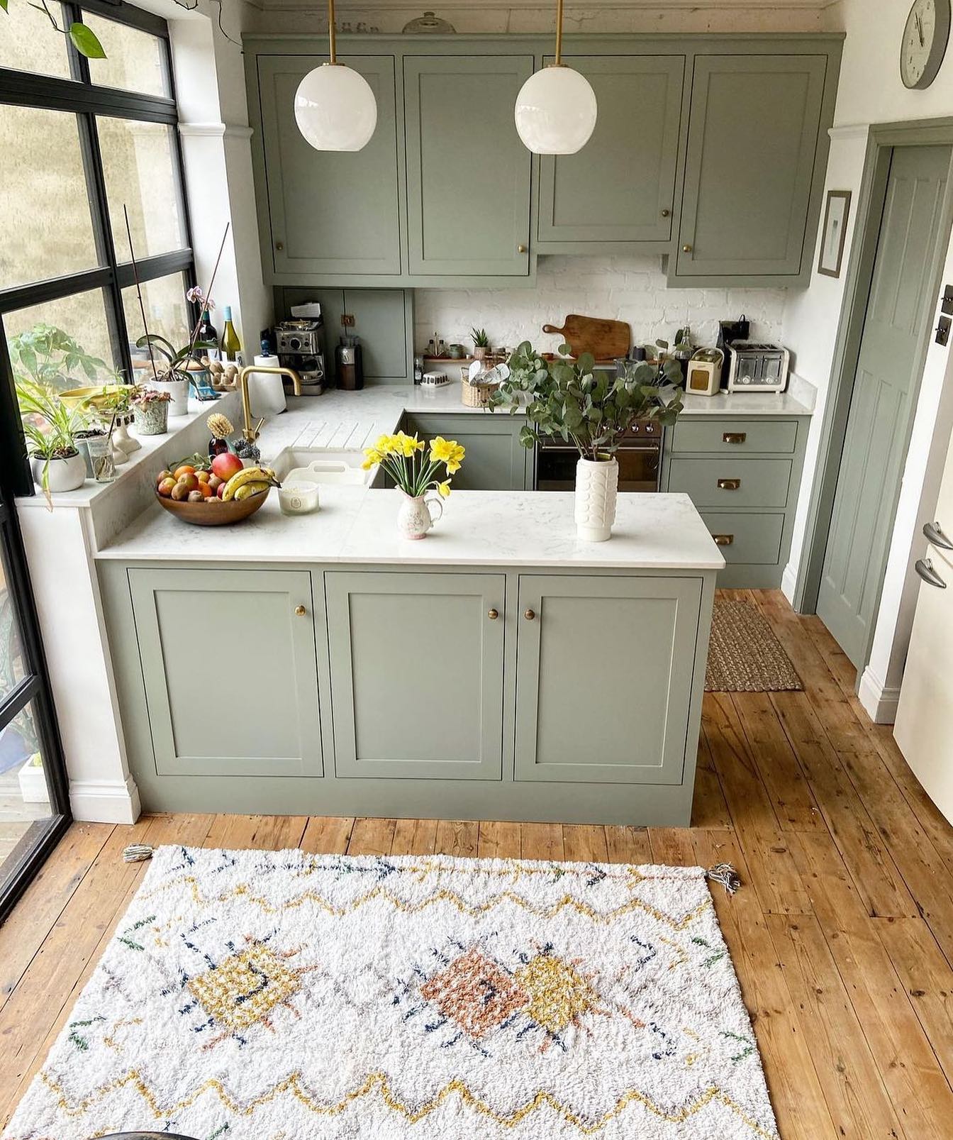 Sage green U-shaped kitchen with two white globe pendants, black-framed Crittall-style doors, plants on the windowsill, a Berber-style zigzag rug, and wide wood plank floors