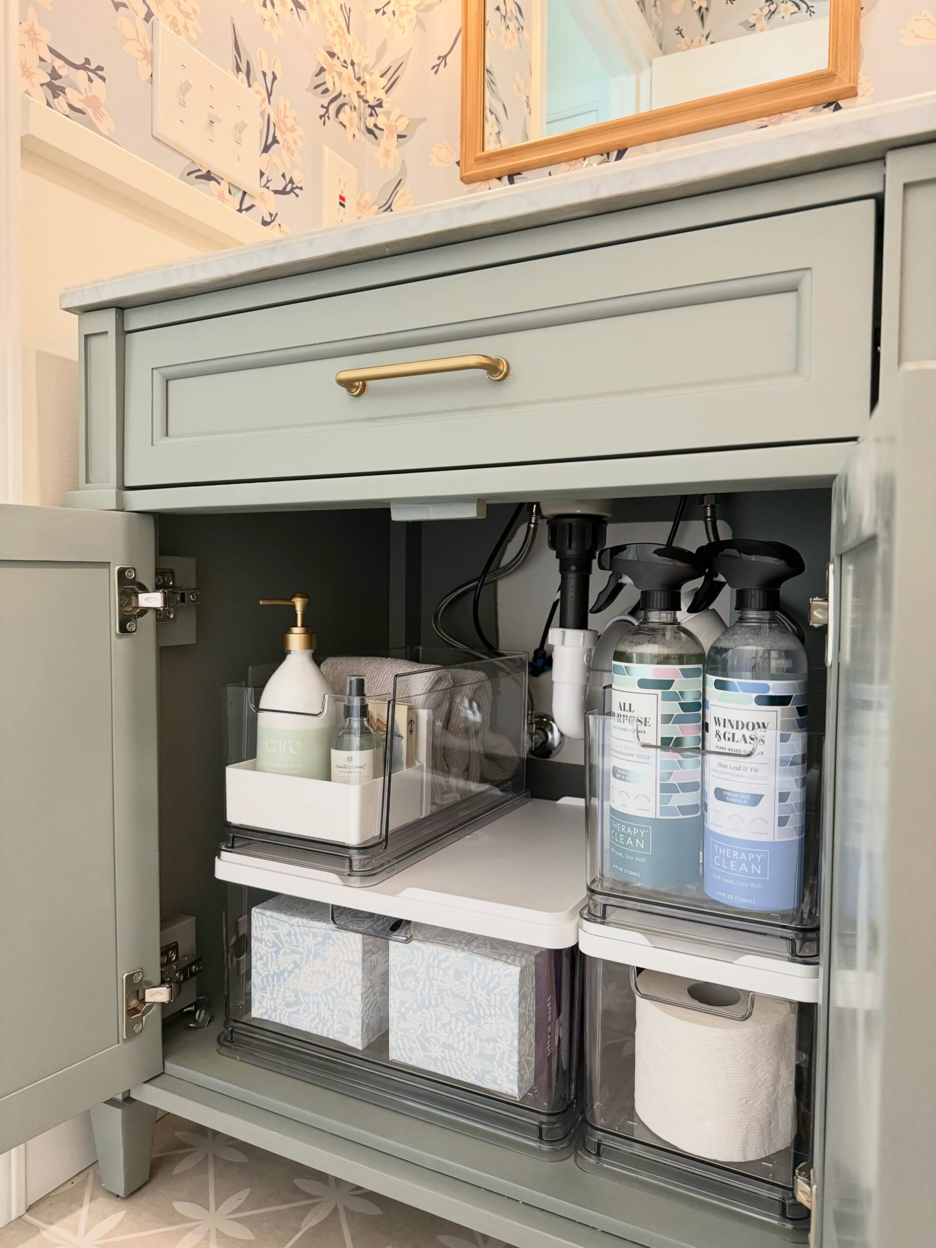 Open sage green bathroom vanity with brass handles showing clear pull-out drawers holding tissue boxes and toilet paper, a smaller caddy with hand lotion and towels on one side, and tall cleaning spray bottles on the other