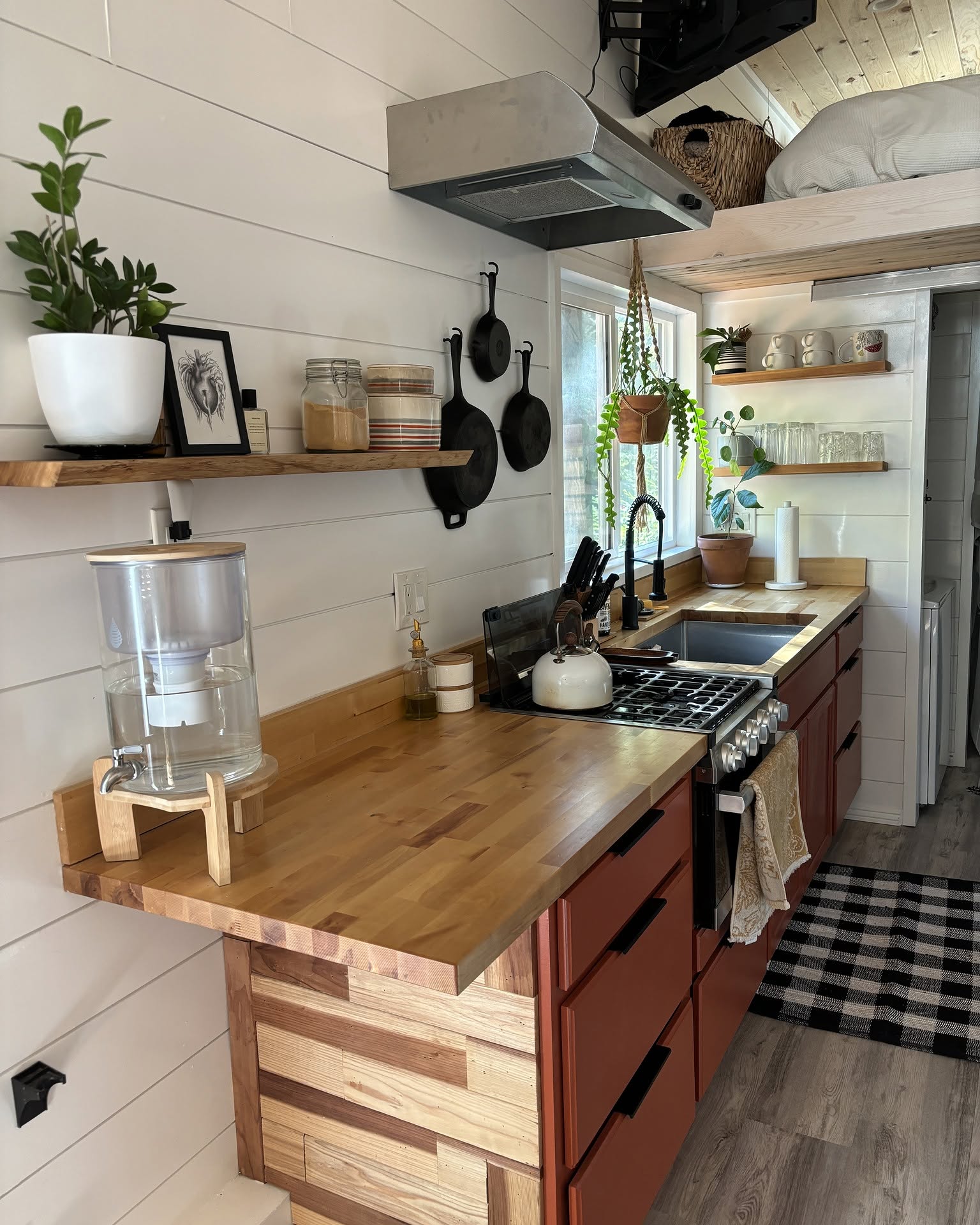 Renovated RV kitchen with sage green lower cabinets, white quartz counter, subway tile backsplash with dark grout, white upper cabinets, a woven roller shade, and a glimpse of the bathroom at the back