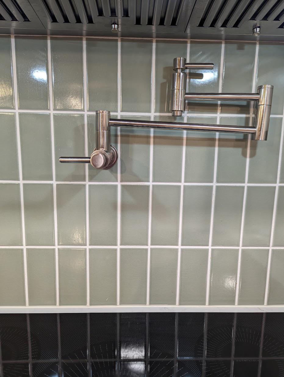 Brushed stainless pot filler mounted on a sage green vertical stack tile wall above a black induction cooktop, with the stainless hood visible at the top of the frame.