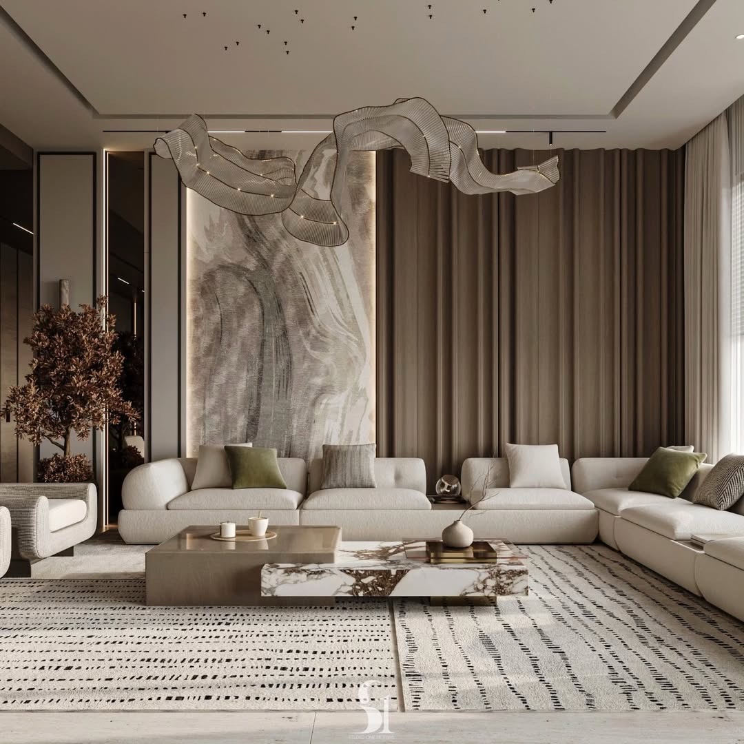 Cream linen sectional sofa with olive green and gray pillows under a sculptural flowing gold ribbon-shaped pendant light, with a large abstract brushstroke painting backlit on the wall behind, dual coffee tables in bronze and white marble, floor-to-ceiling warm linen curtains, and a dark bronze tree sculpture on the left