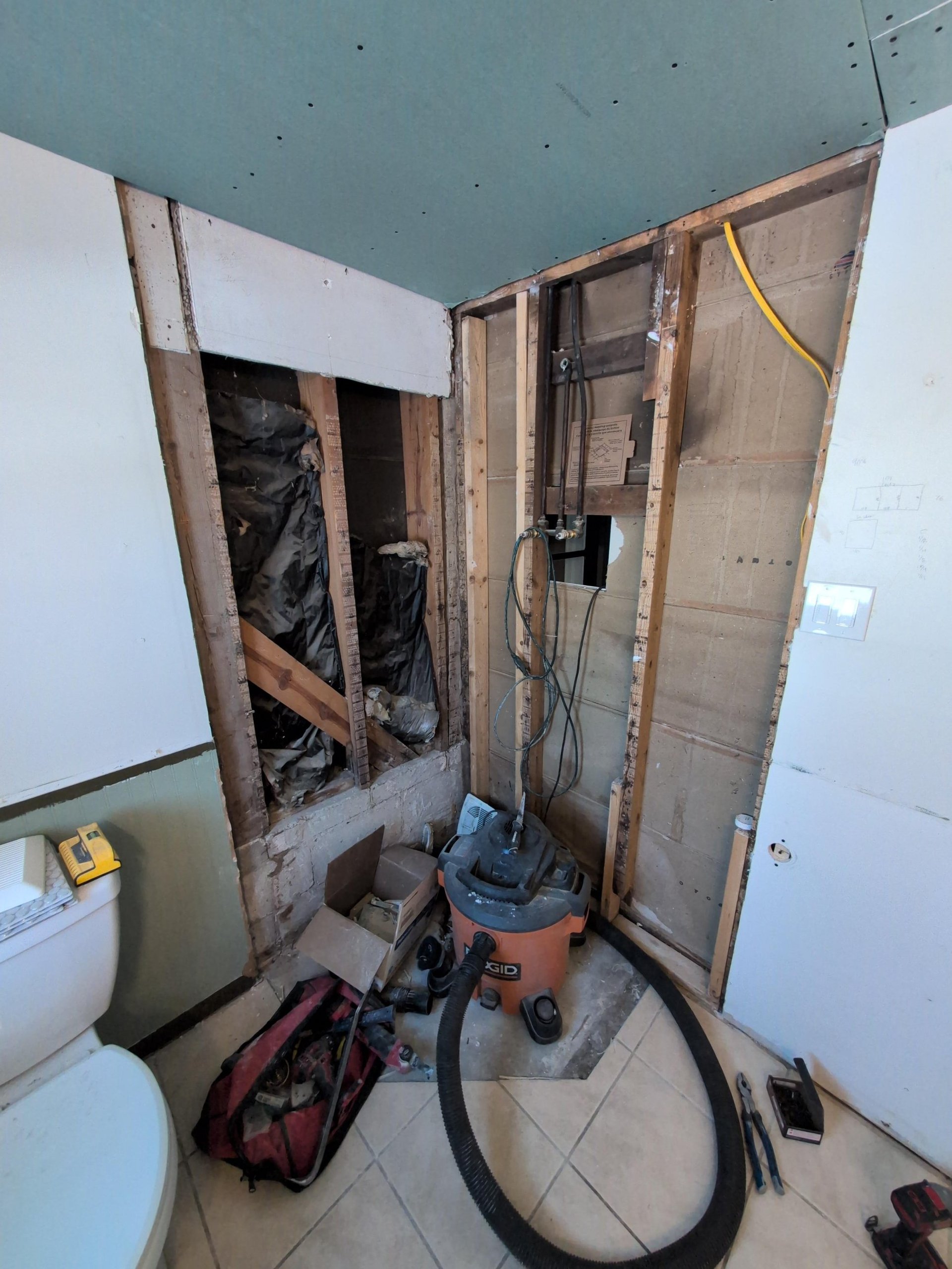 Shower corner torn down to the bare wood studs with the original black vapor barrier torn open, old plumbing lines still in the wall, a Milwaukee tool bag and shop vac on the tile floor, and new green moisture-resistant drywall overhead.