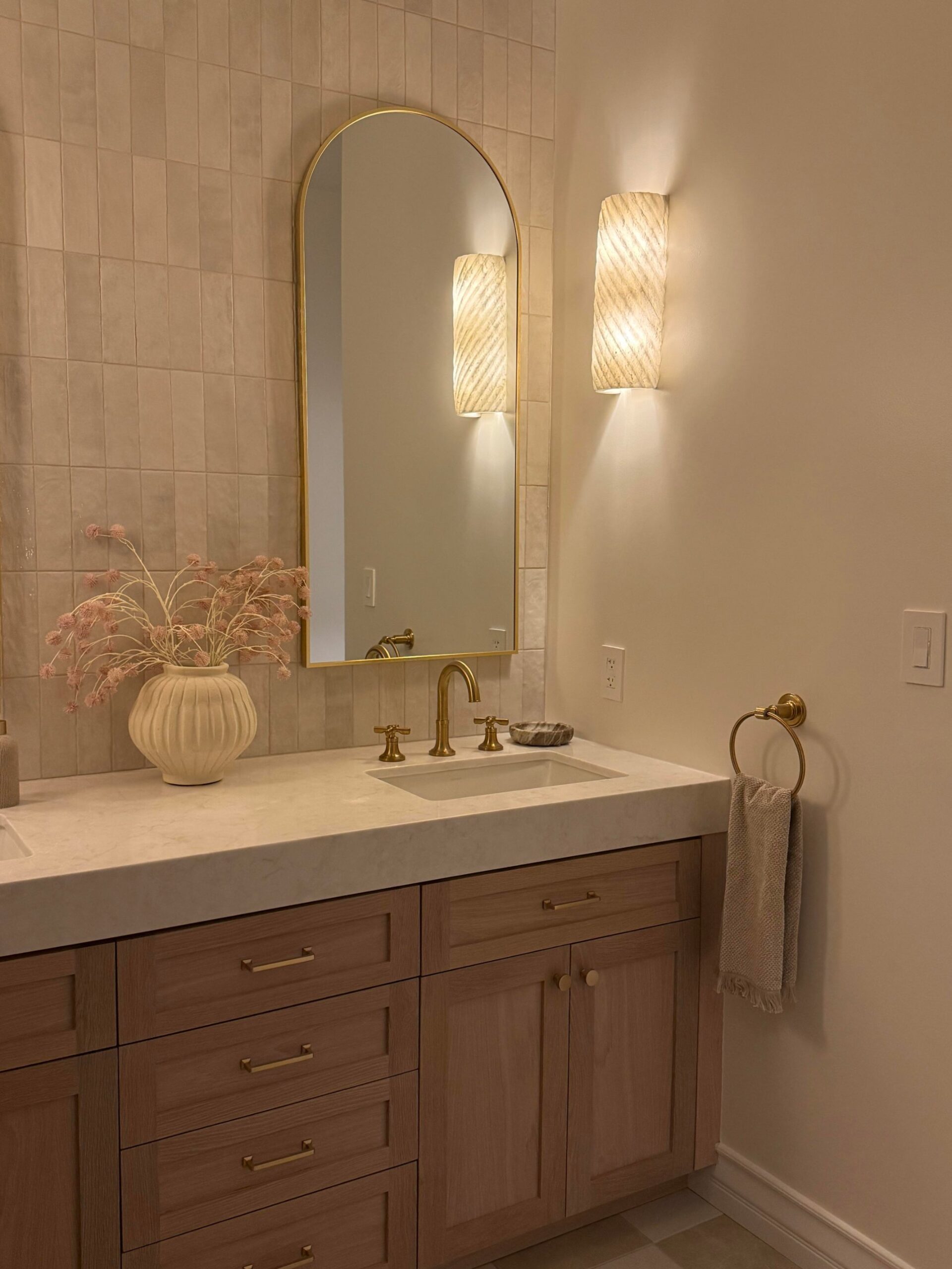 Close-up of one side of the vanity with a tall arched brass mirror, a pleated plaster sconce, white quartz counter, undermount sink, brass gooseneck faucet, cream vase with dried pink flowers, and a brass towel ring.