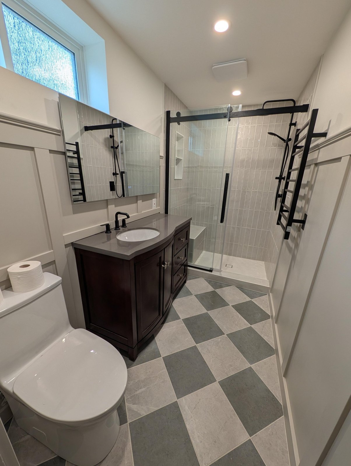 Remodeled bathroom with a gray and cream slate checkerboard floor, dark wood vanity with a gray quartz top, matte black fixtures, a glass sliding shower door, and a tall black heated towel ladder on the wall.