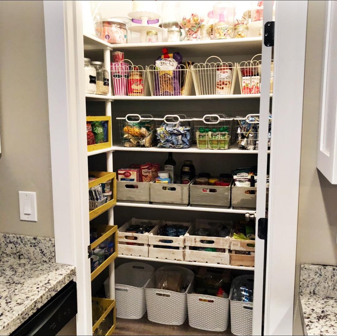 Small white closet pantry with hanging wire baskets on the back wall, whitewashed wooden crates on the middle shelves, white plastic bins on the bottom shelf, and yellow pull-out spice shelves mounted on the side wall