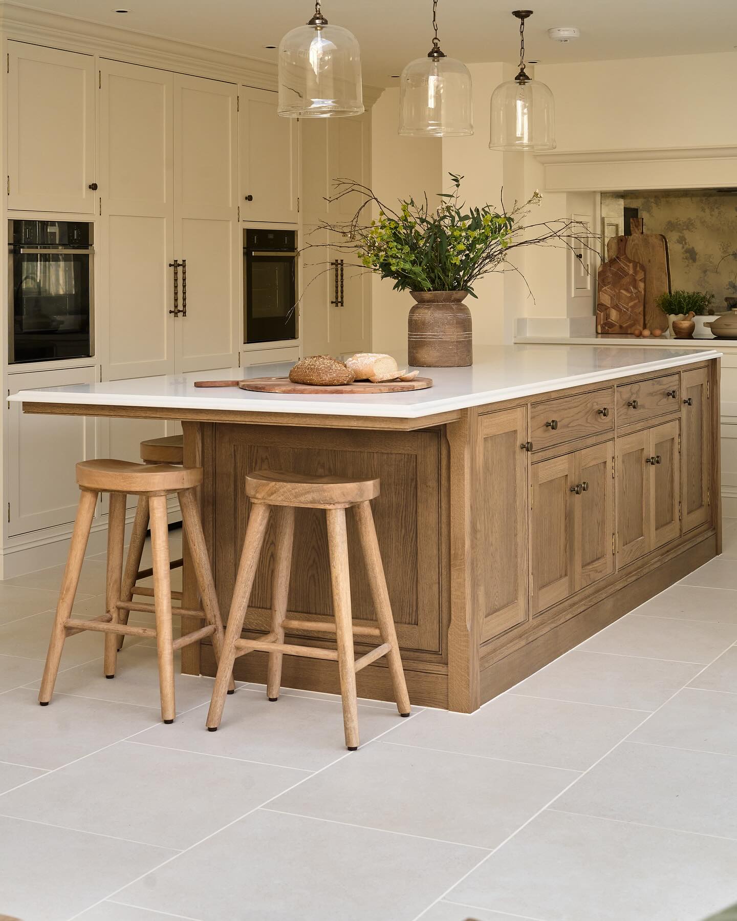 English-style kitchen with a smoked oak island, cream shaker perimeter cabinets, white marble island counter, three clear glass bell pendants, oak bar stools, and a rustic vase with branches