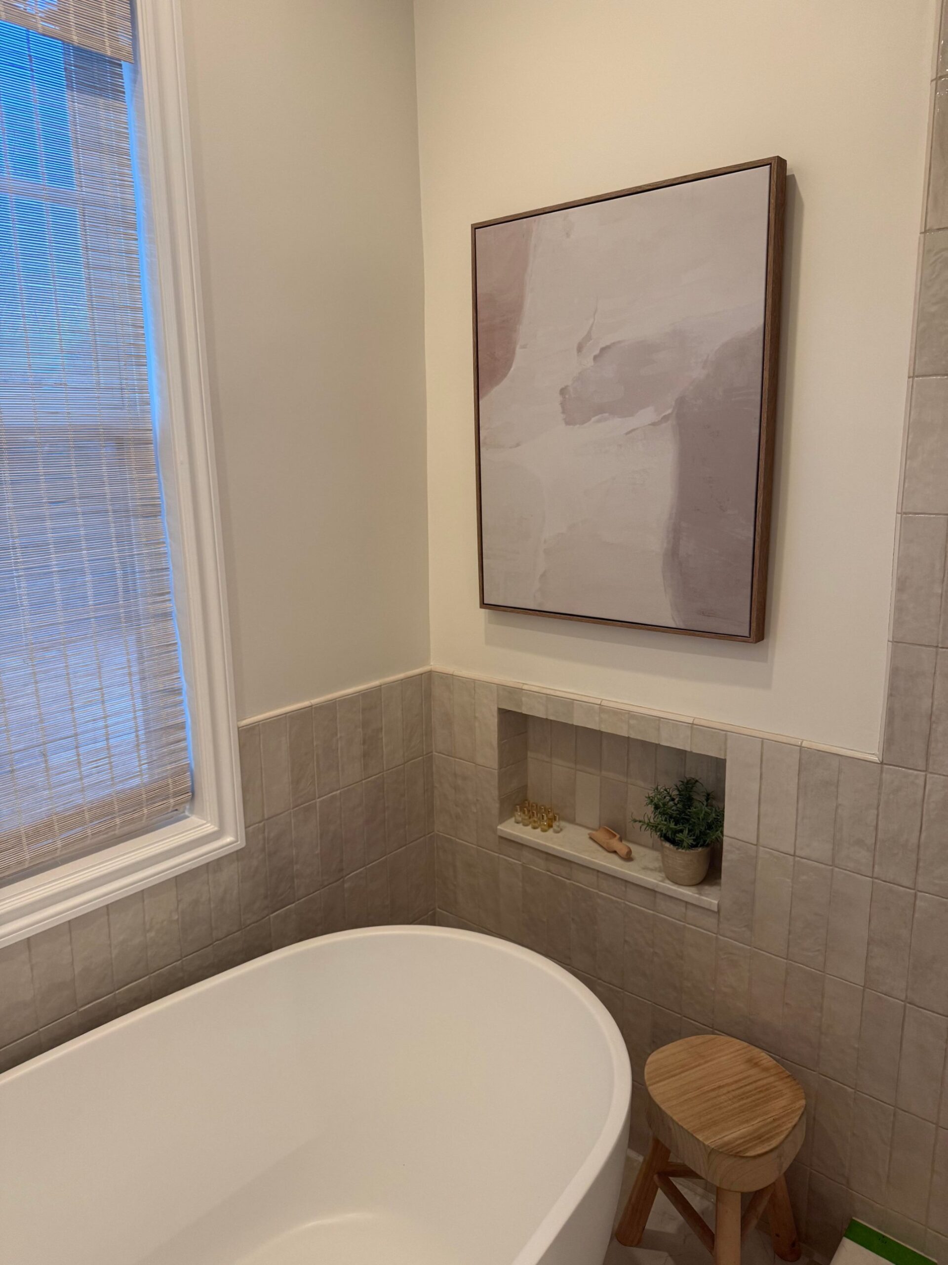 Corner of a bathroom with a white freestanding tub, soft wood-frame abstract art in blush and gray, a built-in tiled niche with marble shelf, a small wooden stool, and a woven window shade.