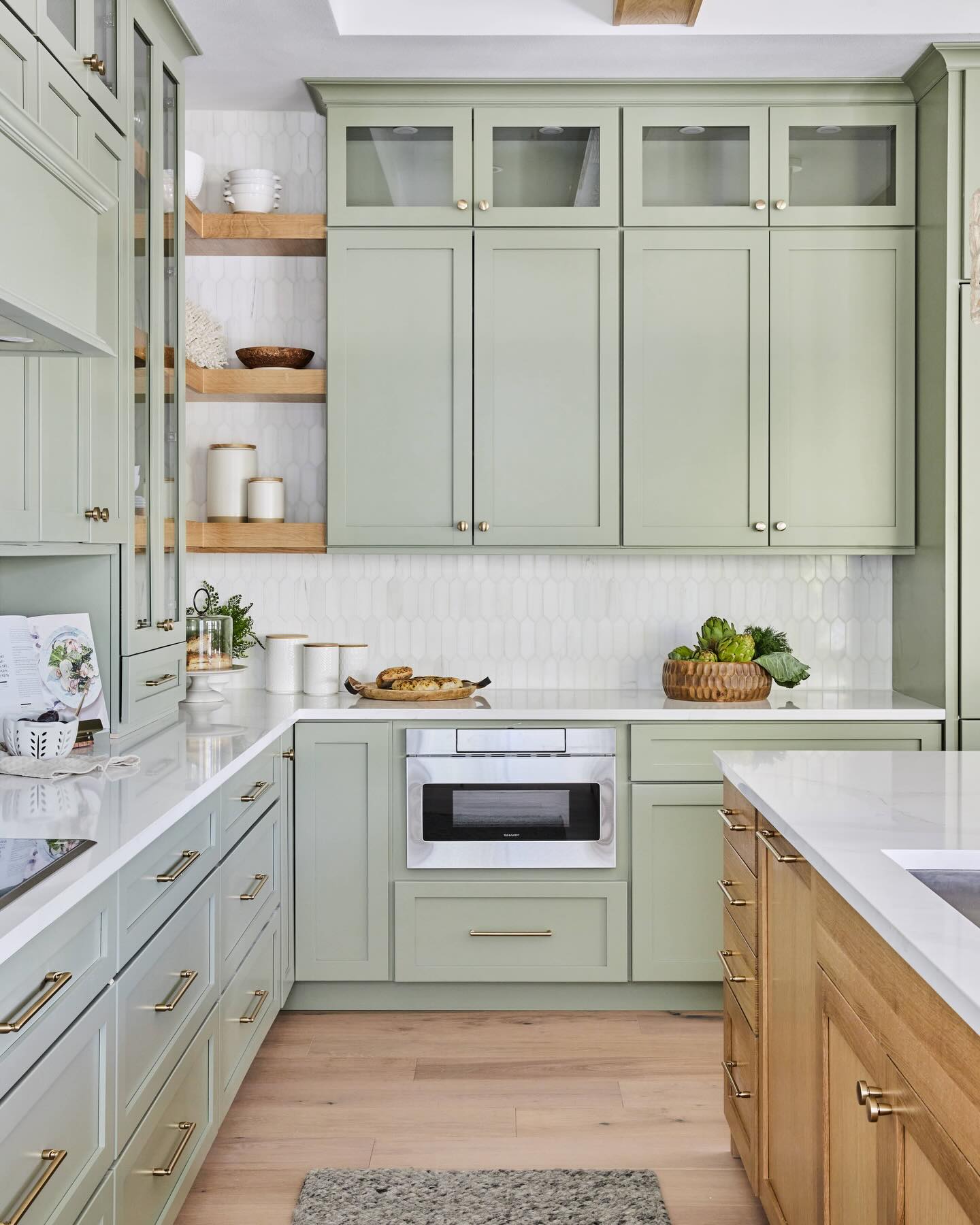 Soft sage green shaker cabinets with gold brass pulls, white picket tile backsplash, floating oak corner shelves, and a wood island on the right