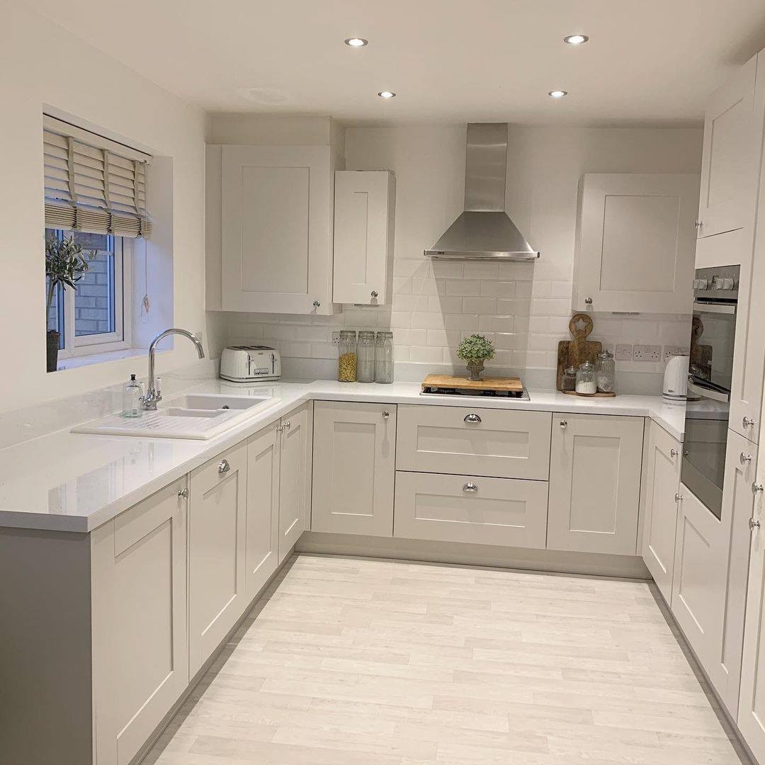 Soft greige U-shape shaker kitchen with white sparkly quartz countertops, stainless steel chimney hood, white subway tile backsplash, chrome cup pulls and round knobs, light tile floor, small window with wooden Roman shade, and white 4-slice toaster on counter