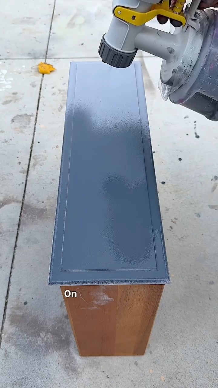 A paint sprayer gun laying down a smooth coat of deep navy blue paint across a primed drawer face lying flat on a concrete driveway.