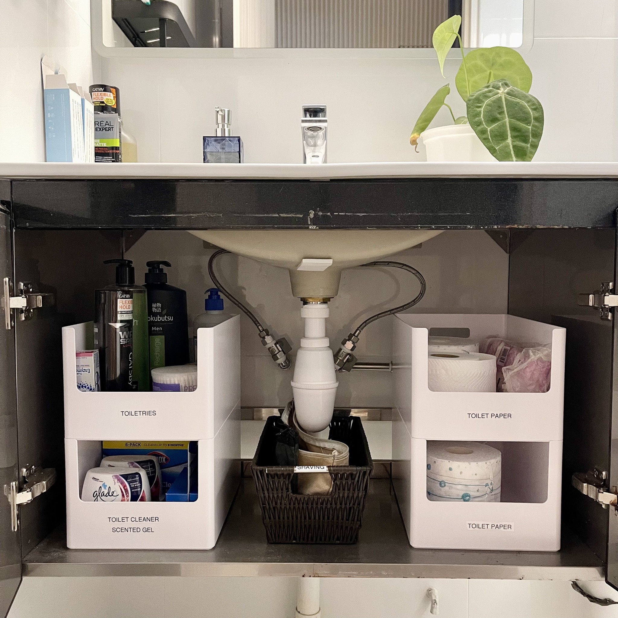 Open black bathroom cabinet with white stackable bins labeled Toiletries, Toilet Cleaner Scented Gel, and Toilet Paper on both sides, with a woven basket labeled Shaving tucked around the central P-trap pipe