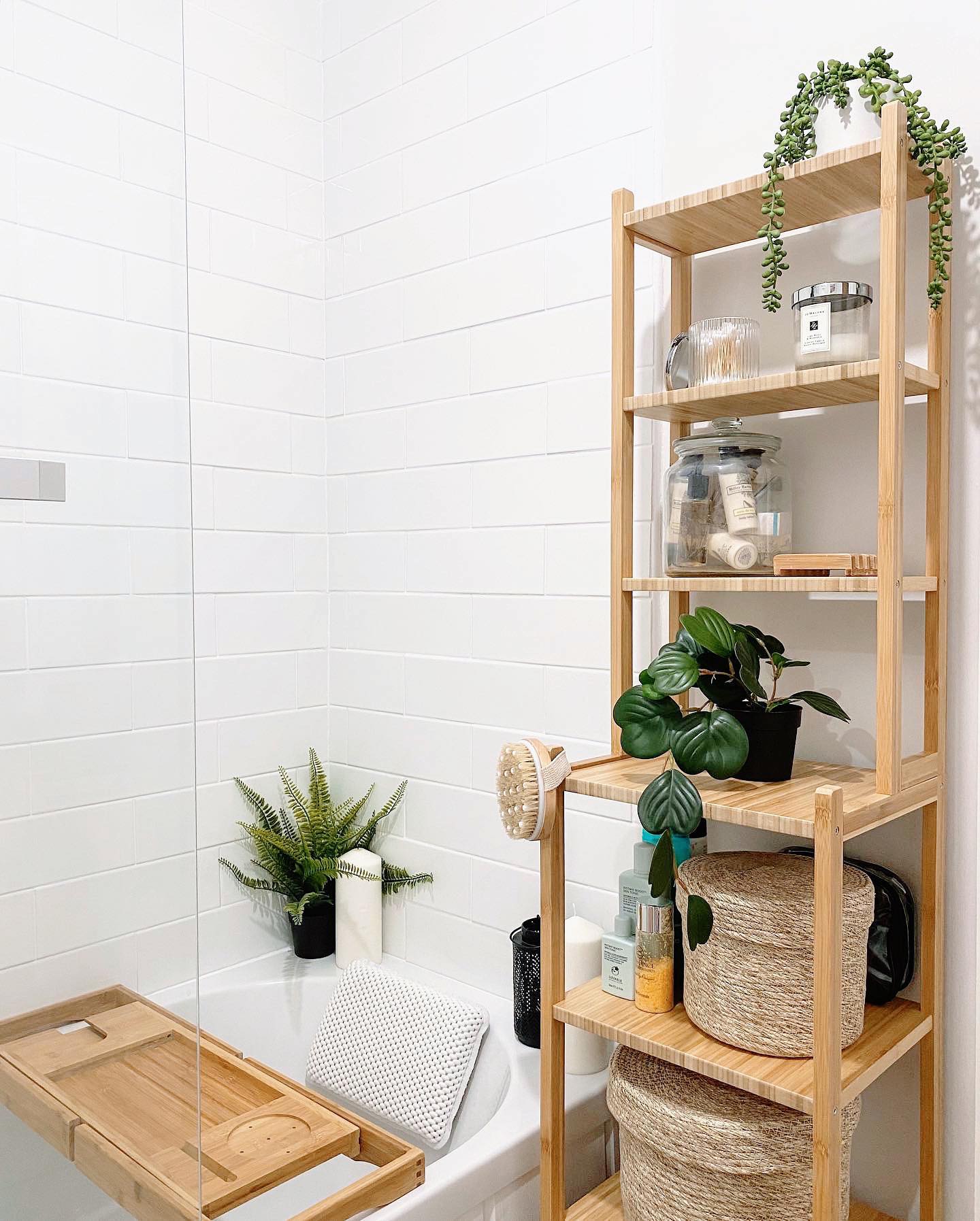 Stepped bamboo shelving unit beside a white tiled bathtub, holding trailing greenery, candles, apothecary jars, a potted plant, and stacked seagrass baskets