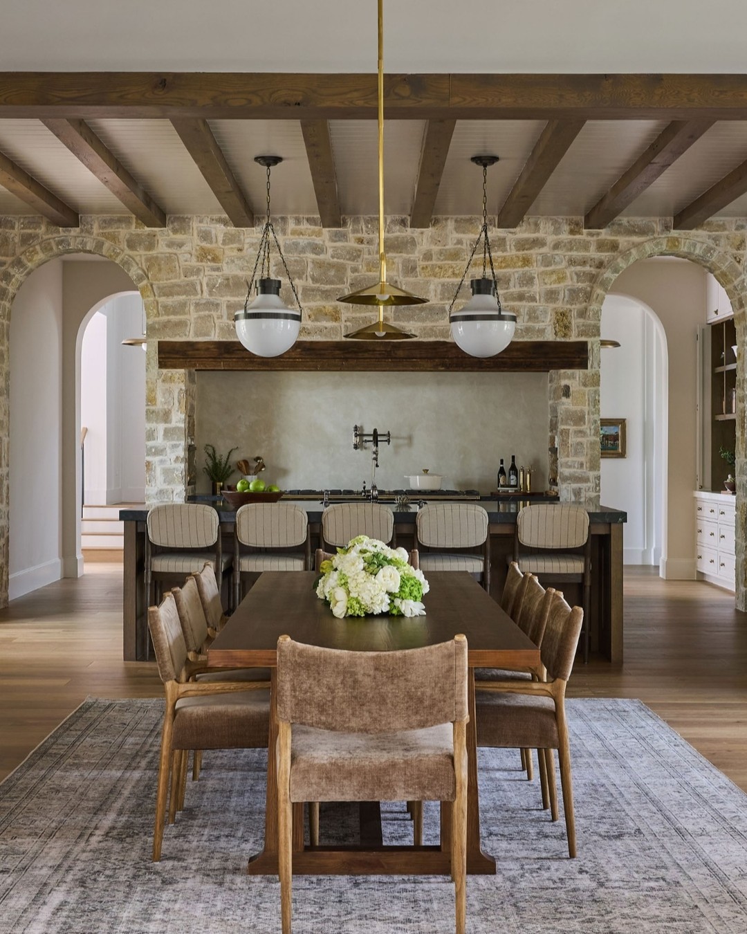 Modern farmhouse dining room with a stone wall and two arched openings, exposed wood ceiling beams, dark wood dining table, upholstered dining chairs, and open kitchen with island in the background