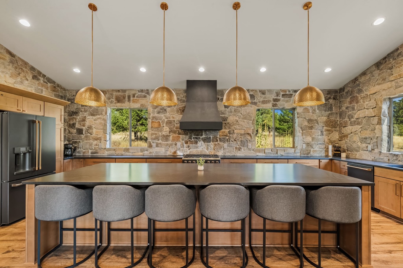 Open cabin kitchen with a full stone accent wall, four hammered brass dome pendants in a row, matte black range hood, warm wood shaker cabinets, large black-topped island, and six gray upholstered counter stools