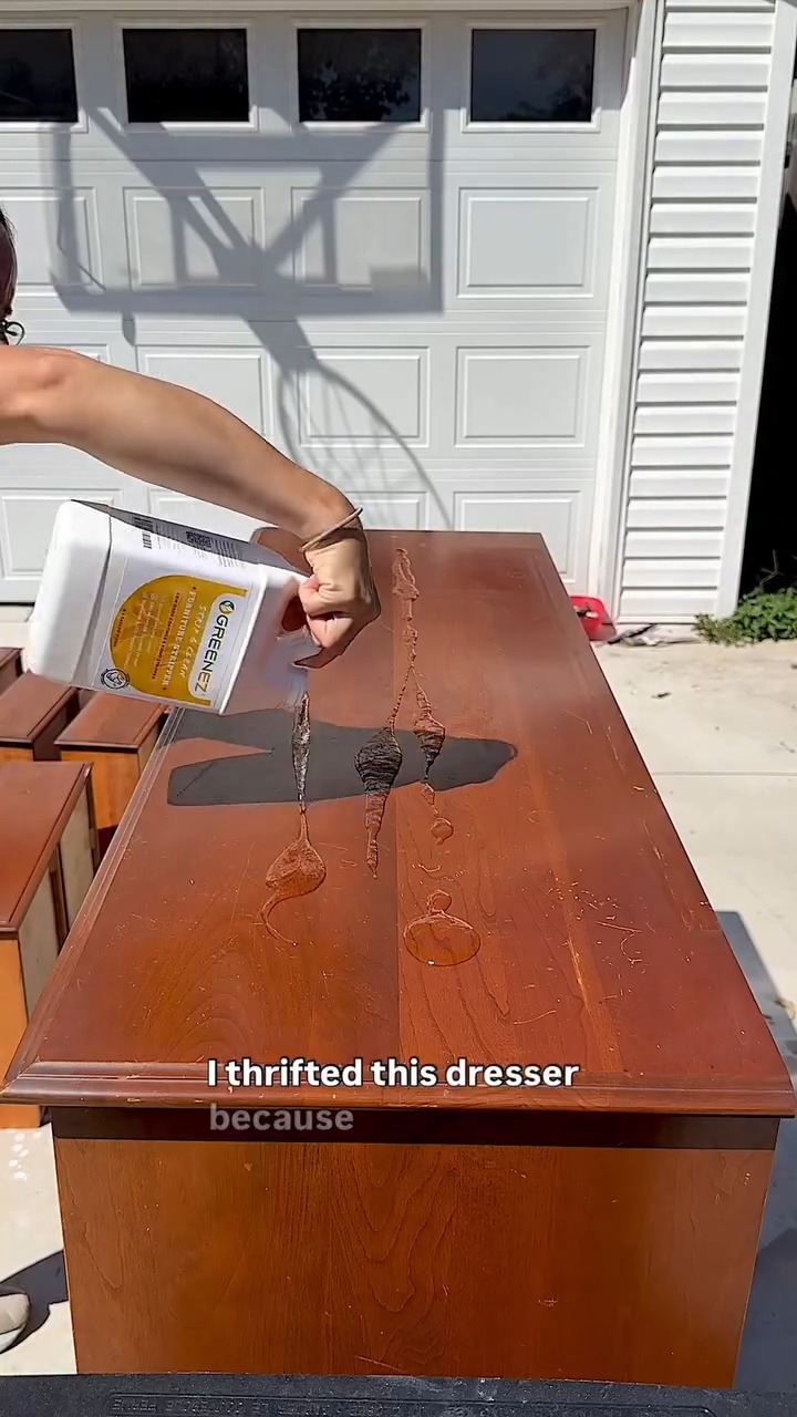 A hand pouring thick chemical stripper from a white Greenez jug onto the cherry-stained top of a dresser, outside in front of a white paneled garage door.