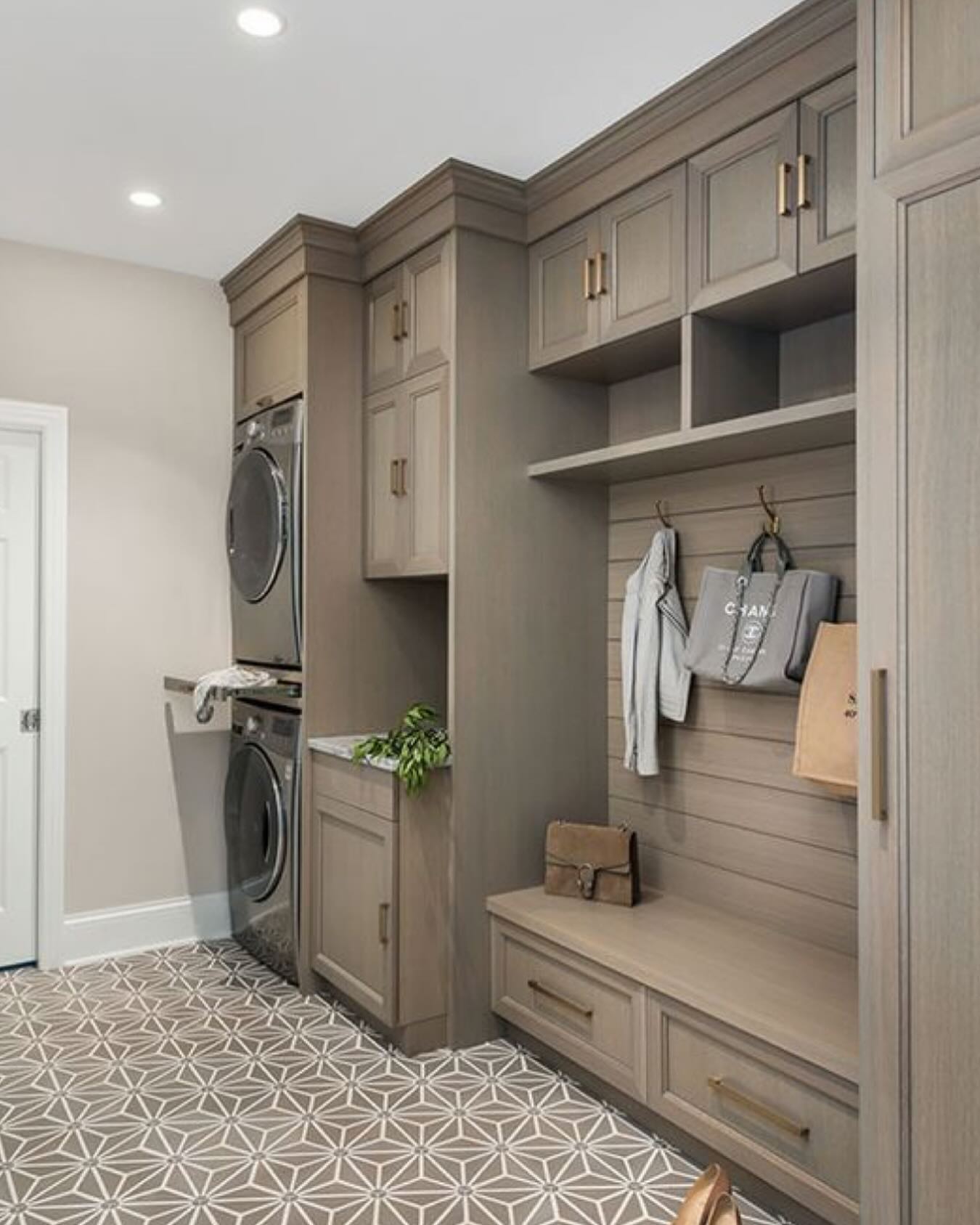 Warm taupe cabinets with brass hardware and horizontal wood plank backing, stacked black washer and dryer, mudroom bench with hooks, Chanel tote and brown suede bag, and a geometric gray and cream star pattern tile floor