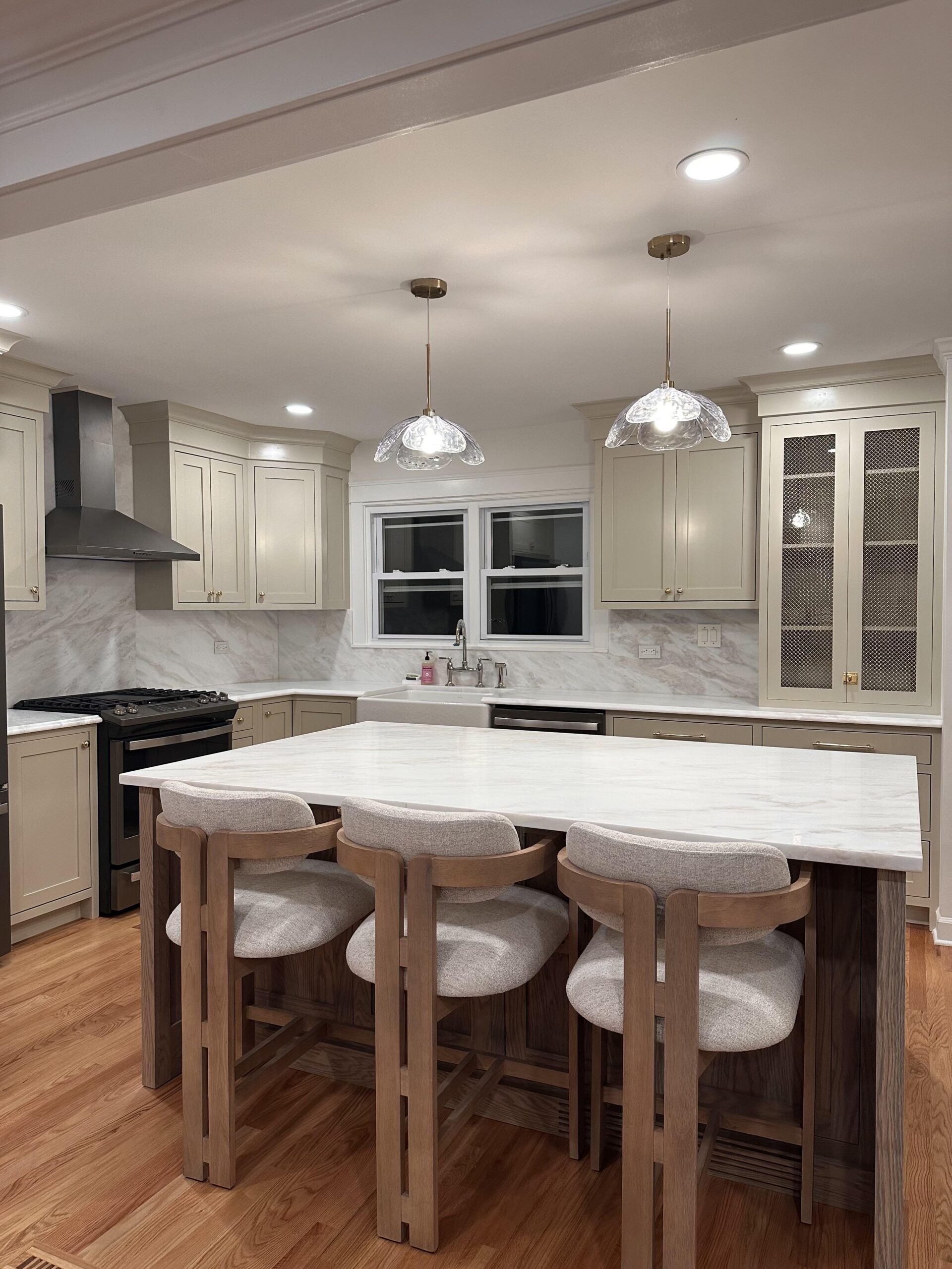 Remodeled kitchen with warm greige perimeter cabinets, a stained wood island topped with white marble, brass hardware, and clear scalloped glass pendants.